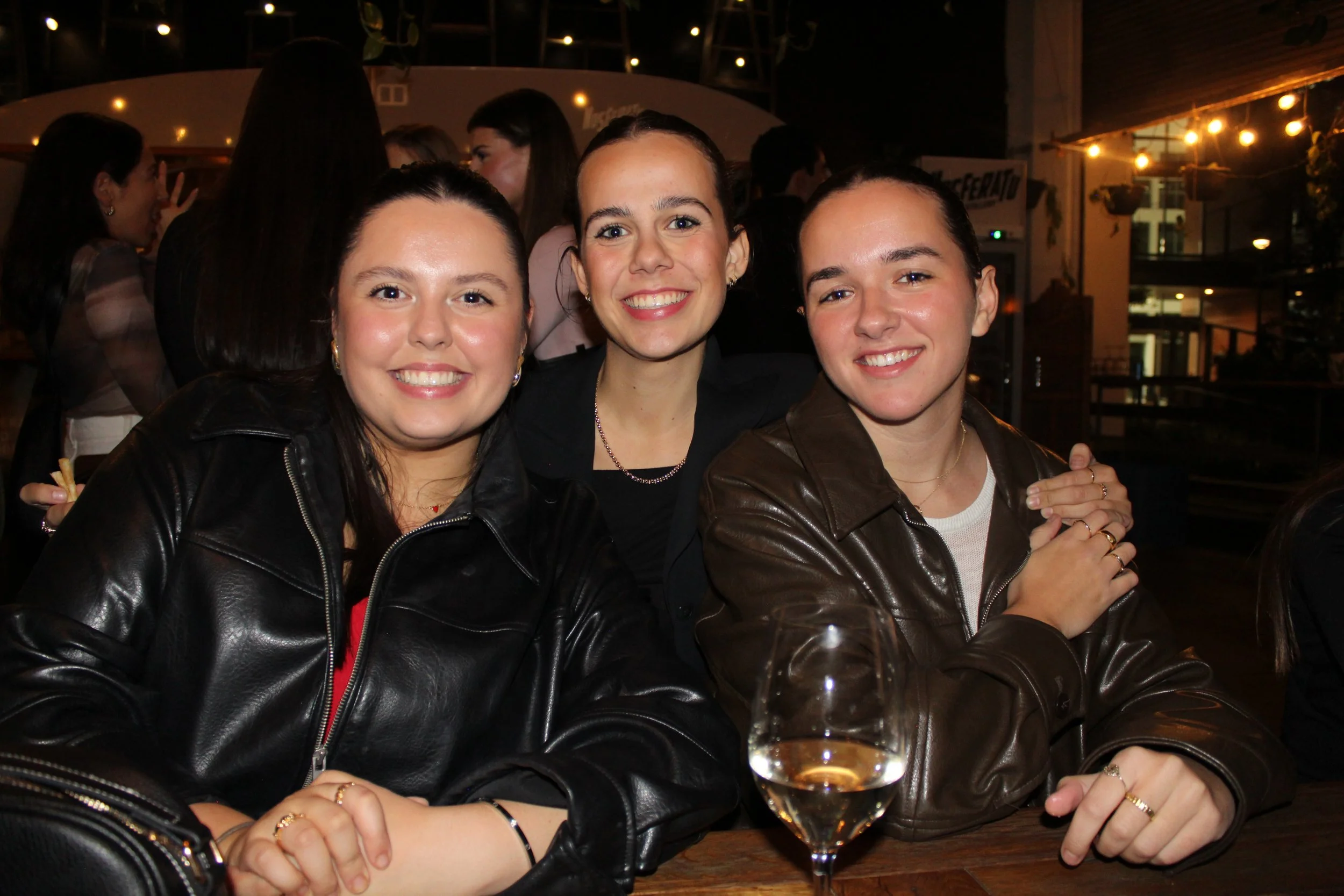 Three women sitting at a table in a dimly lit outdoor setting, smiling at the camera. Two women are wearing leather jackets, and there is a glass of rosé wine in front of them.
