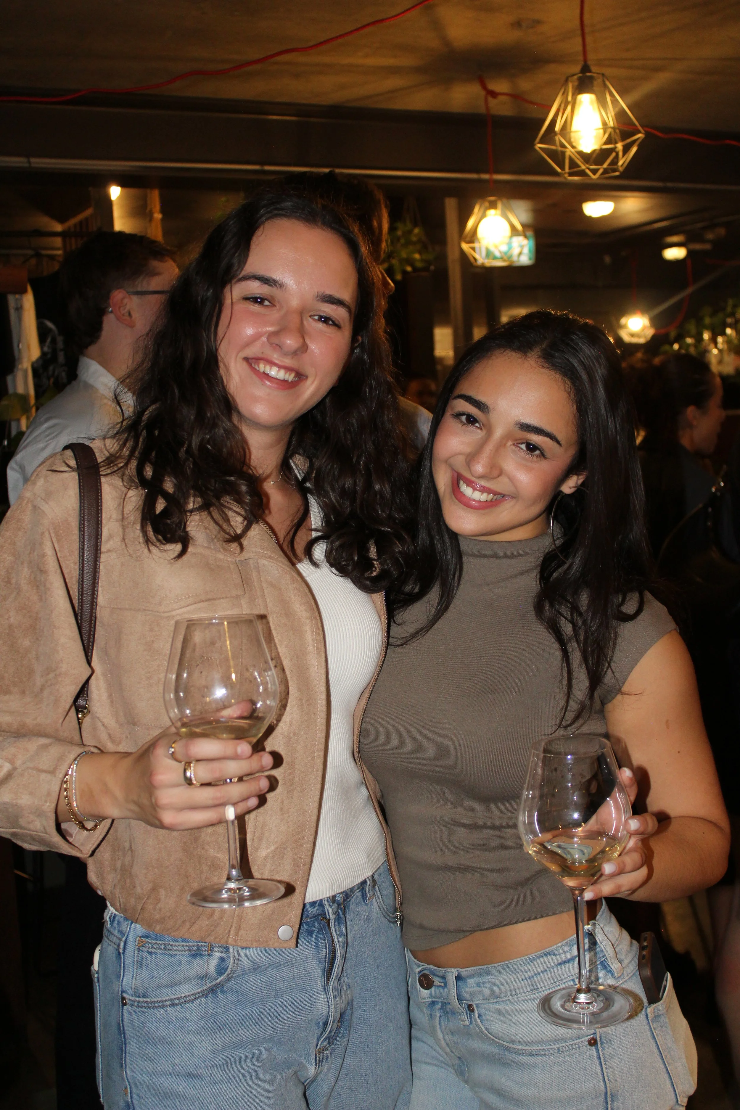 Two women smiling at a social event, holding wine glasses, indoors with warm lighting and modern decor.