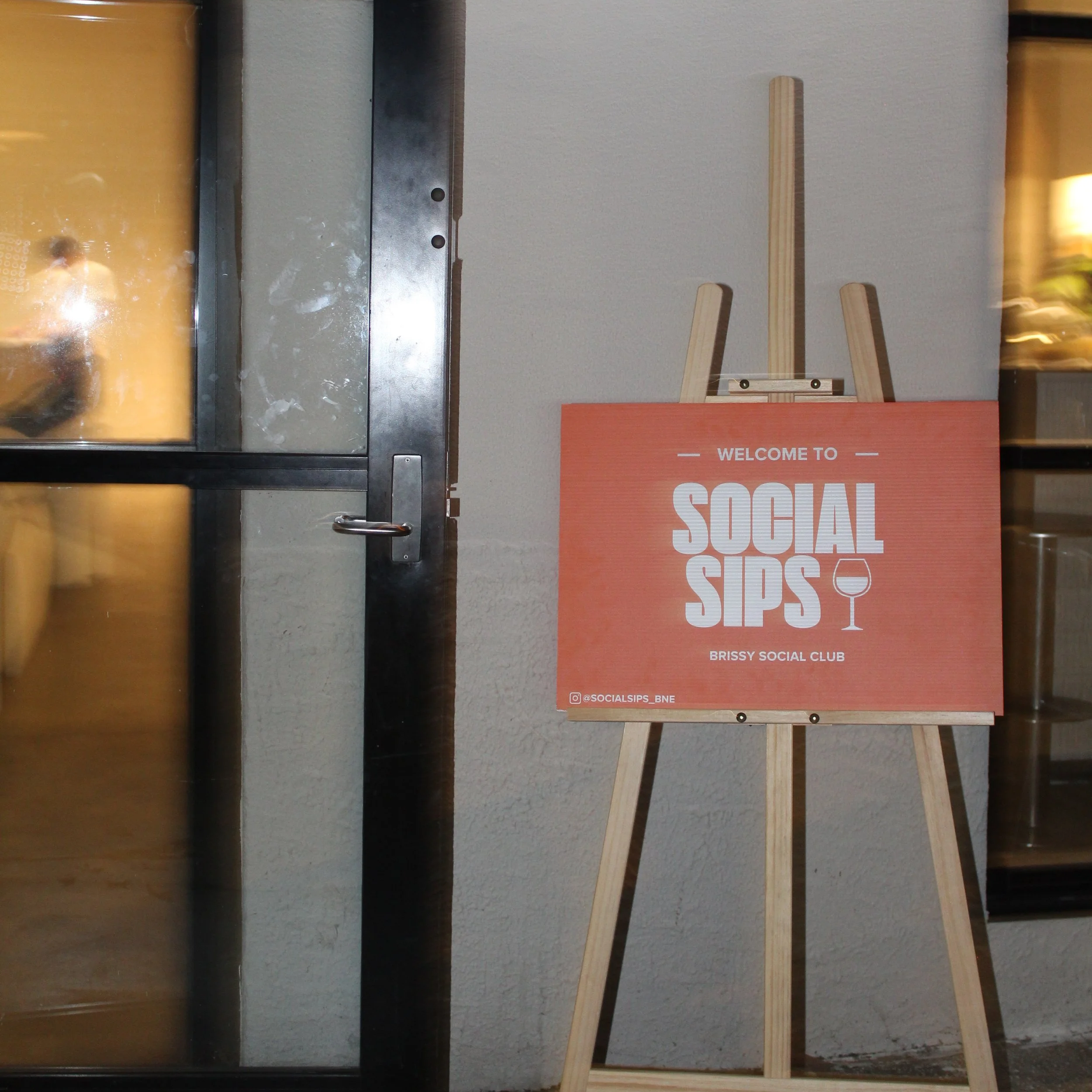 A sign on an easel outside a building reads "Welcome to Social Sips" with a wine glass icon and the Instagram handle @socialsips_bne, indicating an event or venue at Brissy Social Club.