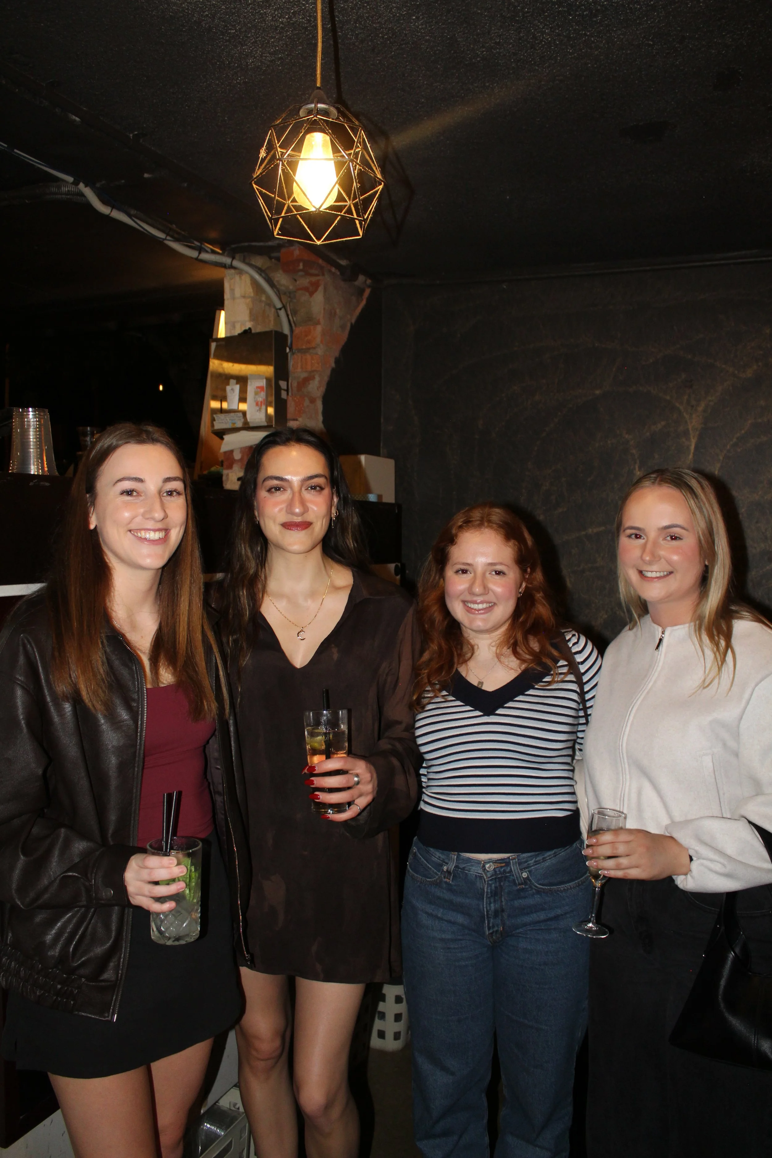 Four women standing together in a bar, holding drinks, smiling for the camera, with a black wall and exposed brick in the background.