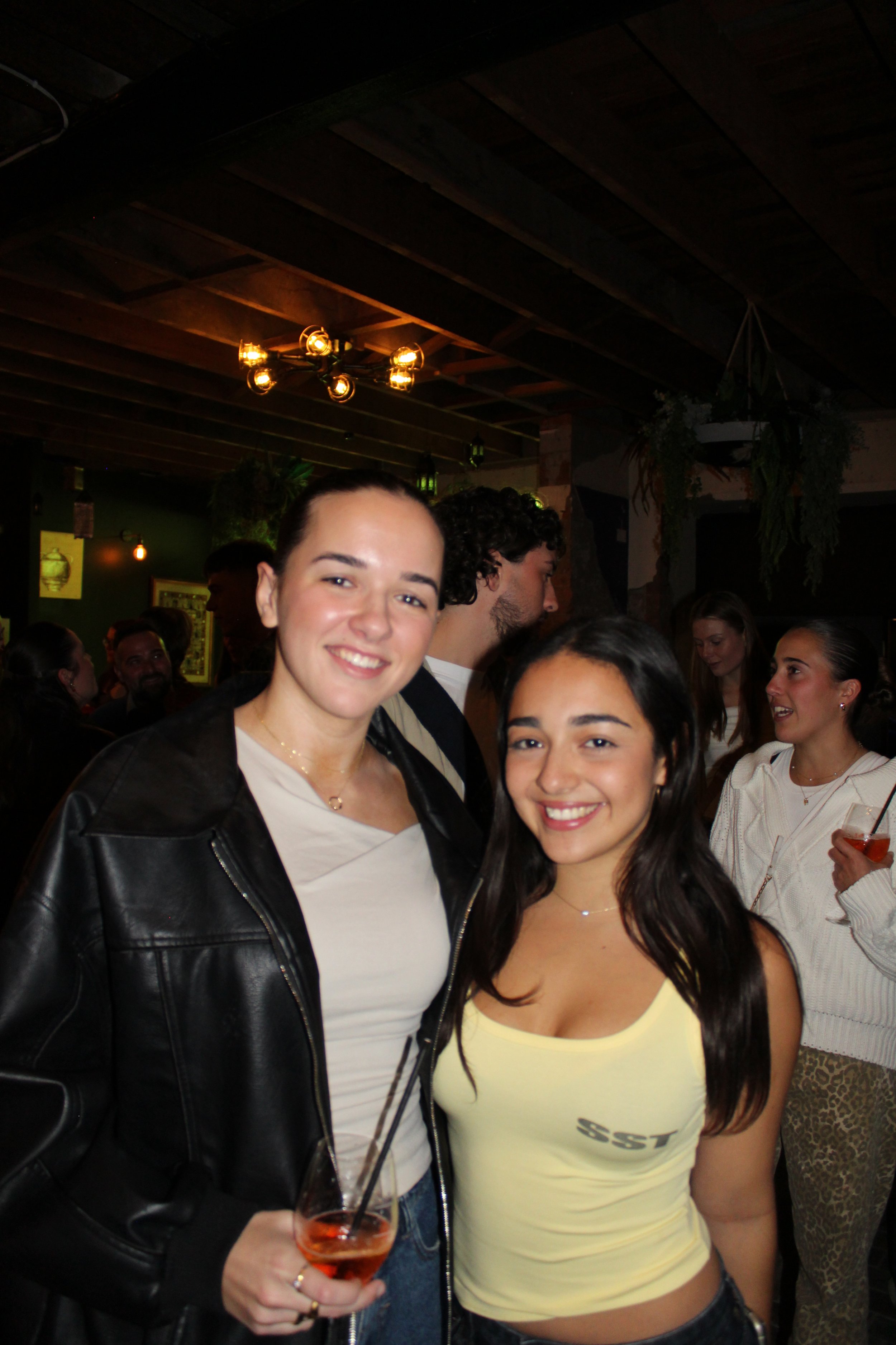 Two young women smiling at a crowded indoor party, one holding a cocktail.