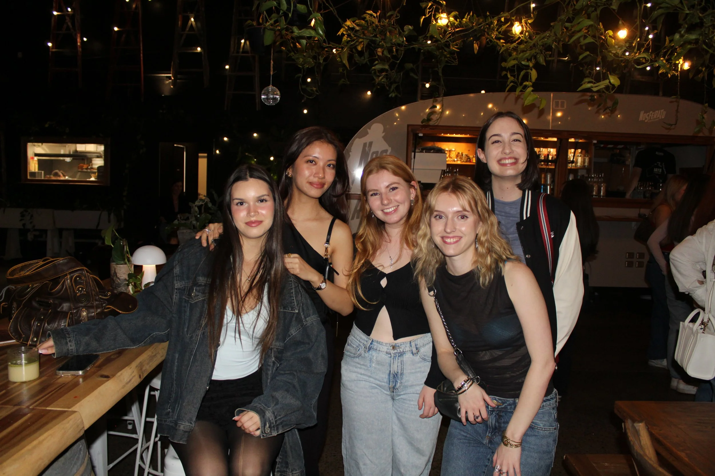 Five women smiling at a night gathering, standing close together in a decorated indoor venue with string lights and plants.