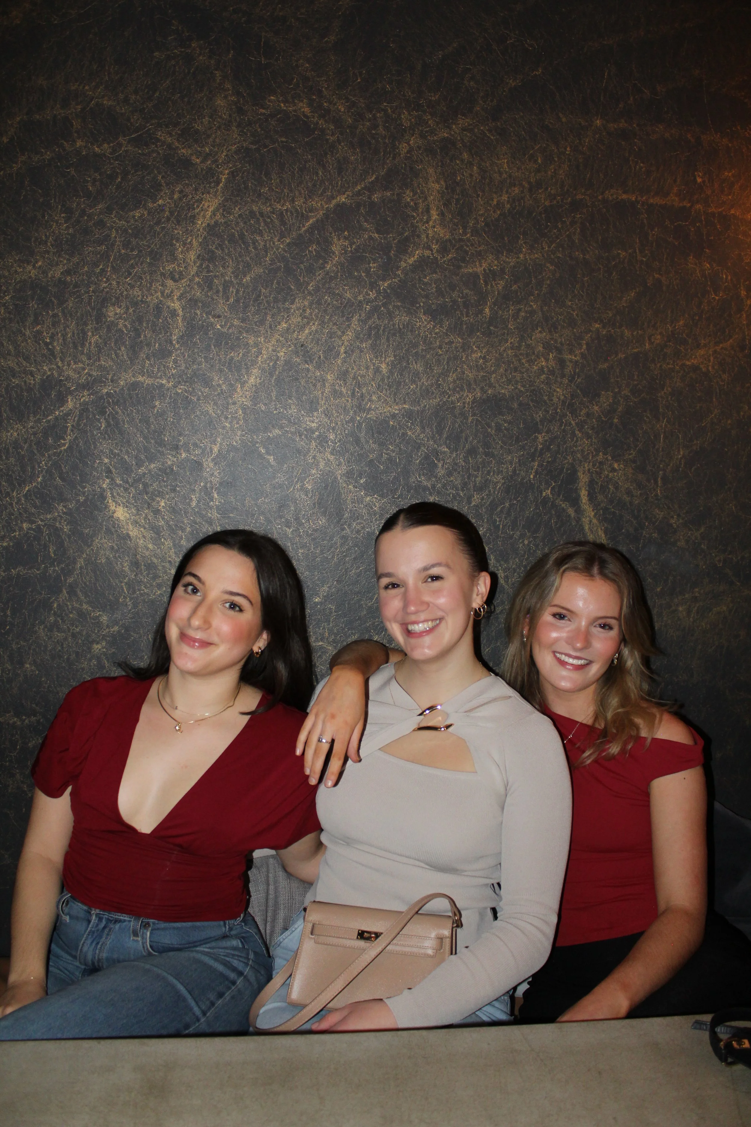 A group of four women posing together, smiling, against a dark, textured background. They are dressed in casual and semi-formal clothing.