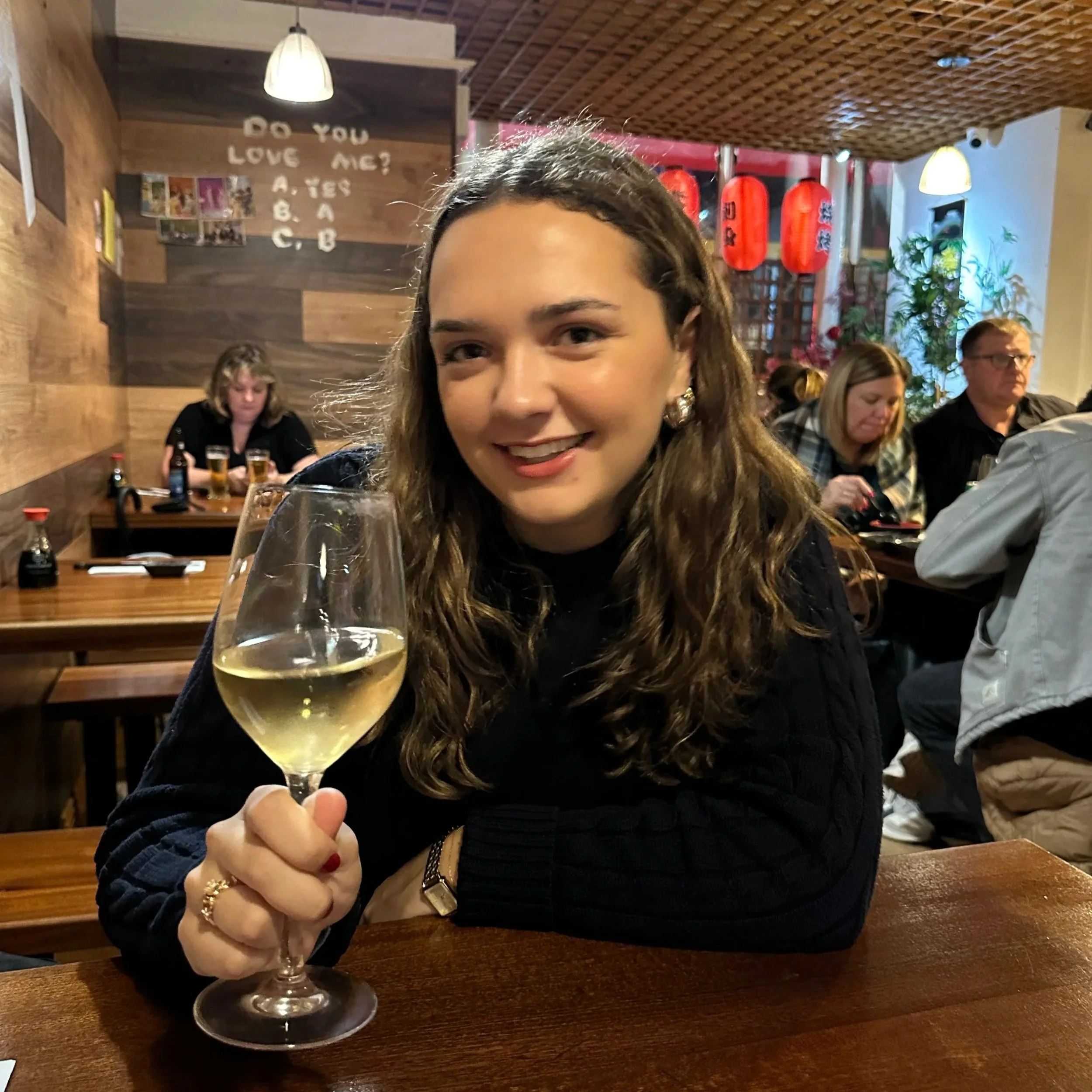 A woman with long curly brown hair and earrings holding a glass of white wine, seated at a wooden table in a restaurant with other diners and Japanese lantern decorations in the background.