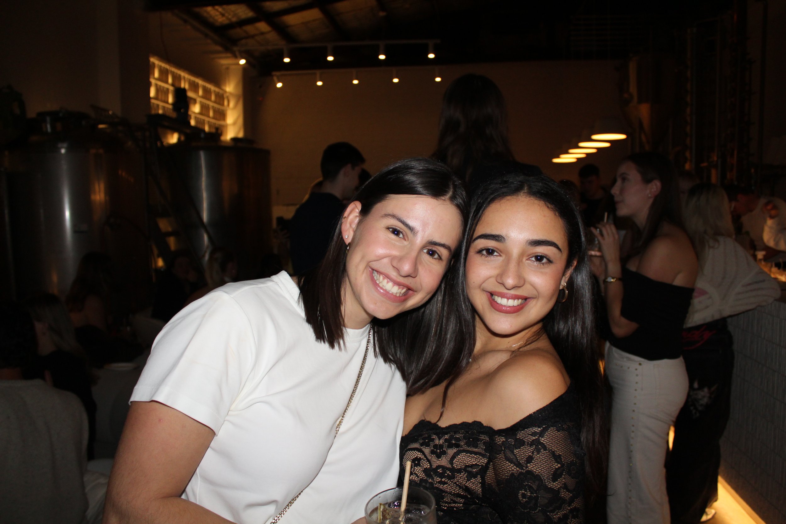 Two women smiling at a social gathering, one wearing a white top and the other wearing a black lace top, with a drink in a glass in front of them, in a dimly lit indoor venue with other people in the background.