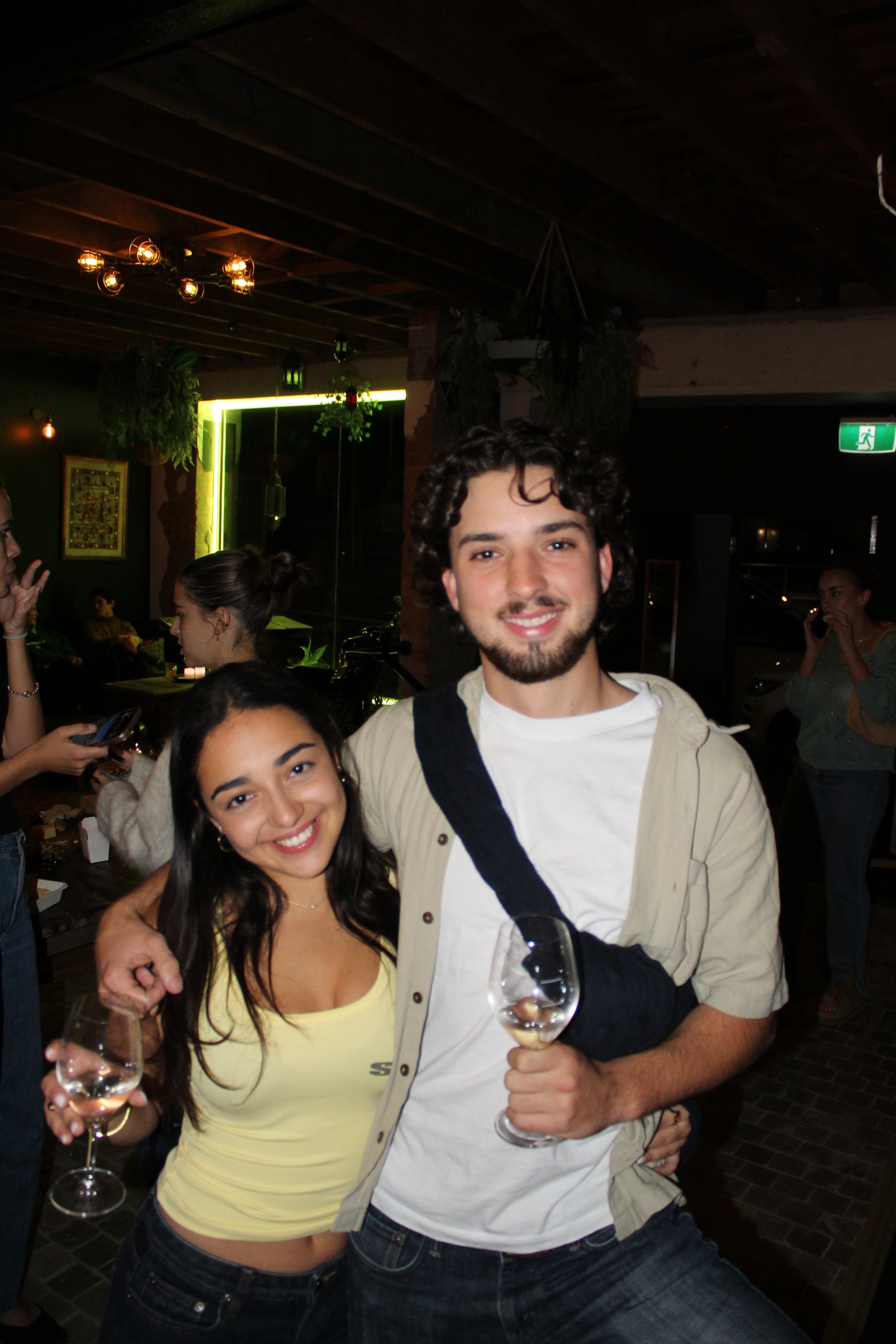 Smiling young woman and man holding wine glasses in a dimly lit bar or restaurant, background with hanging plants and people socializing.