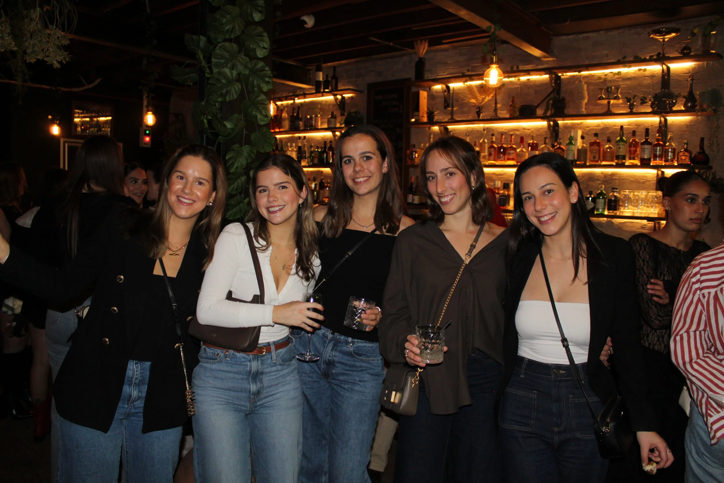 Group of six young women smiling at a bar or club with dark interior, warm lighting, and liquor bottles on shelves behind them.