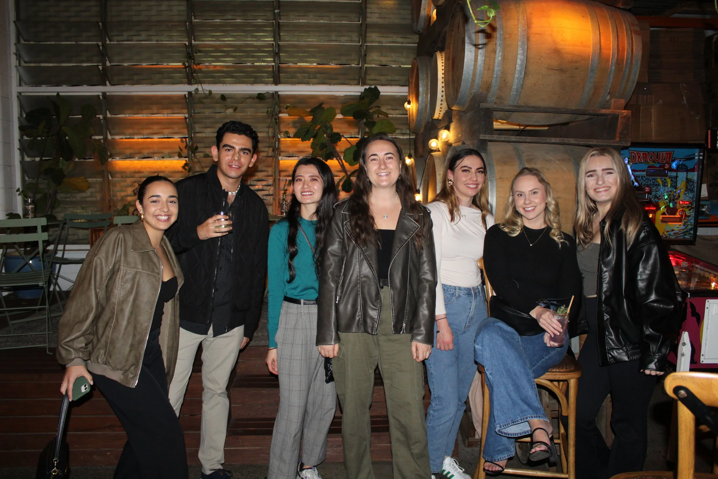 Group of seven young adults smiling at a social gathering in an indoor setting with wine barrels and greenery in the background.