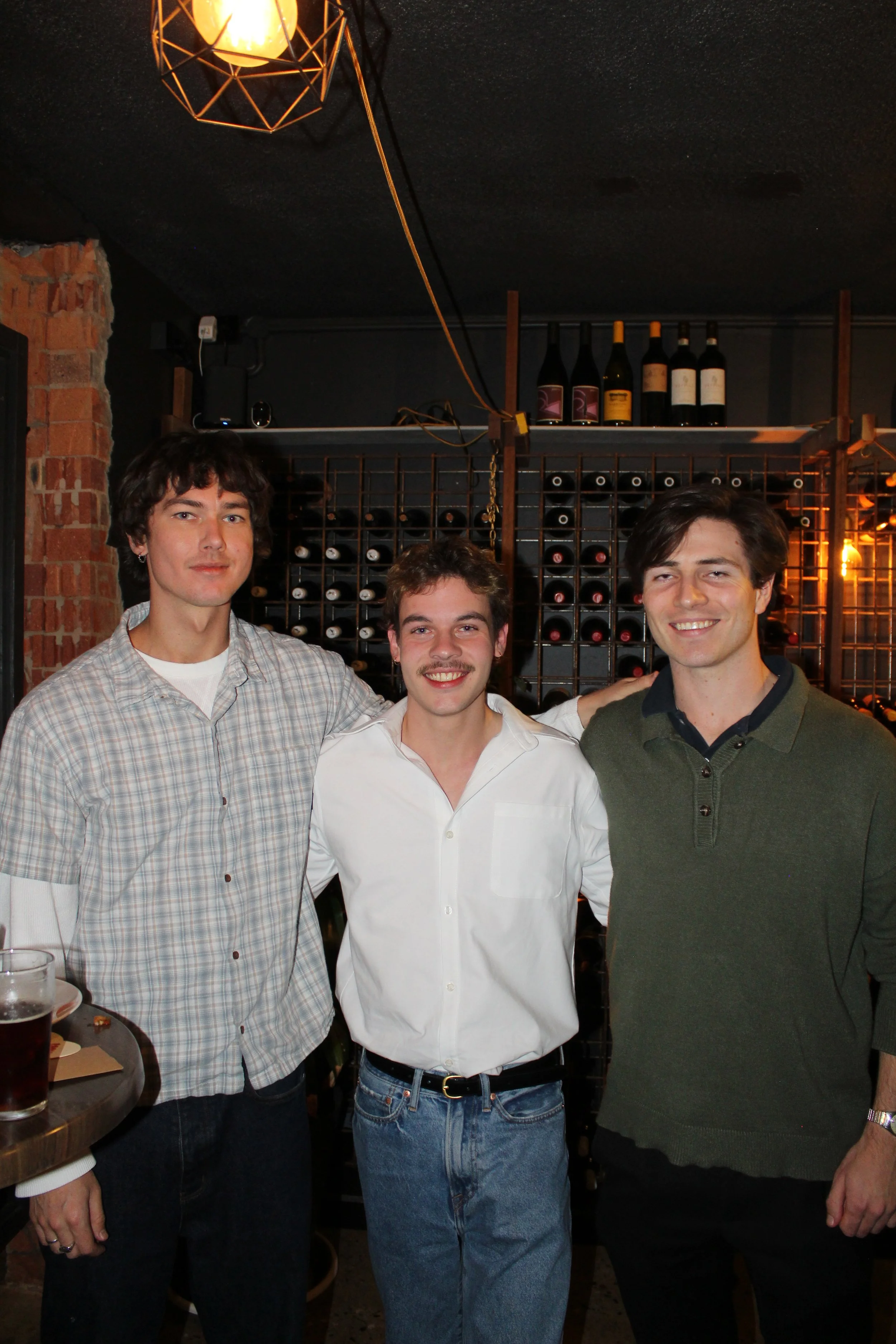 Three young men standing closely together in a dimly lit wine cellar or bar, smiling at the camera. The man on the left wears a plaid shirt, the man in the middle wears a white shirt, and the man on the right wears a green sweater with buttons. Behin