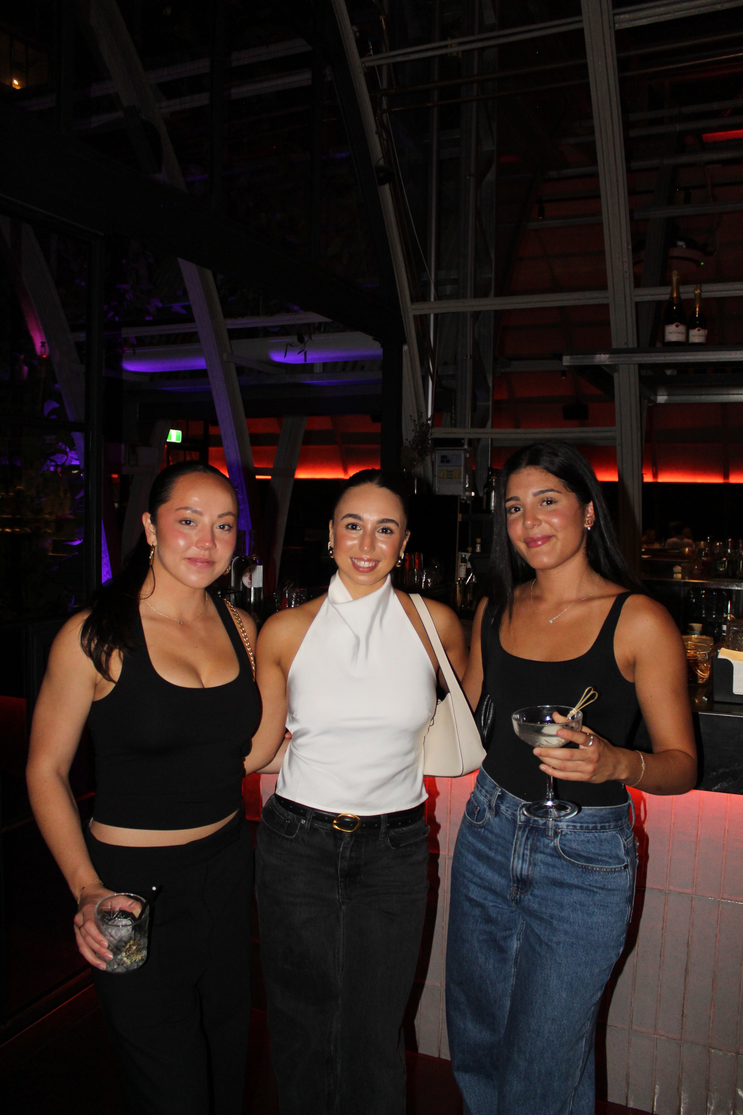 Three women at a bar or nightclub, smiling for the camera, holding drinks, with colorful ambient lighting in the background.