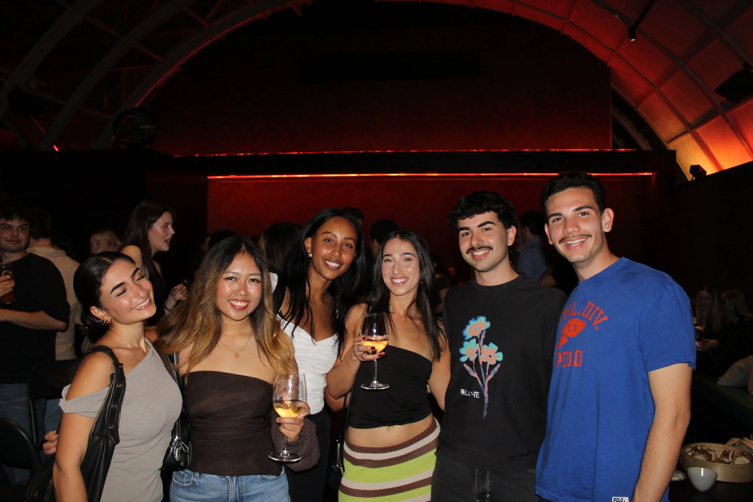 Group of six young adults smiling and holding drinks at a party or social gathering indoors.