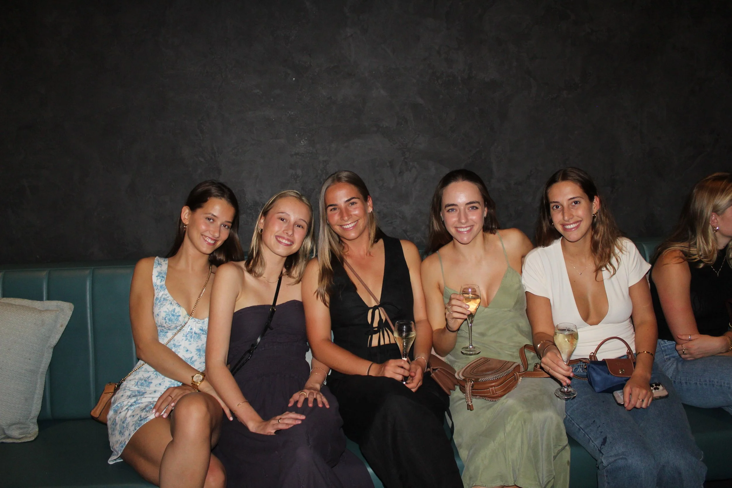 Six women sitting on a green bench against a dark wall, smiling and holding glasses of champagne at a social gathering.