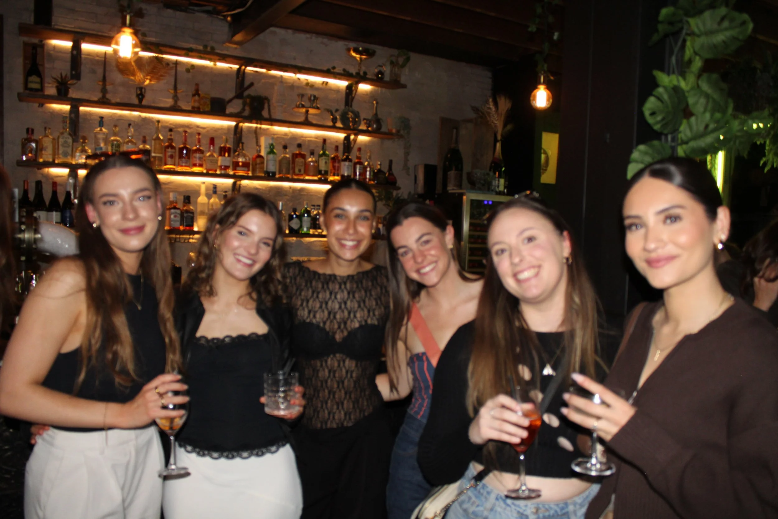 Six women smiling and posing together in a bar, holding drinks, with liquor bottles and shelves in the background.