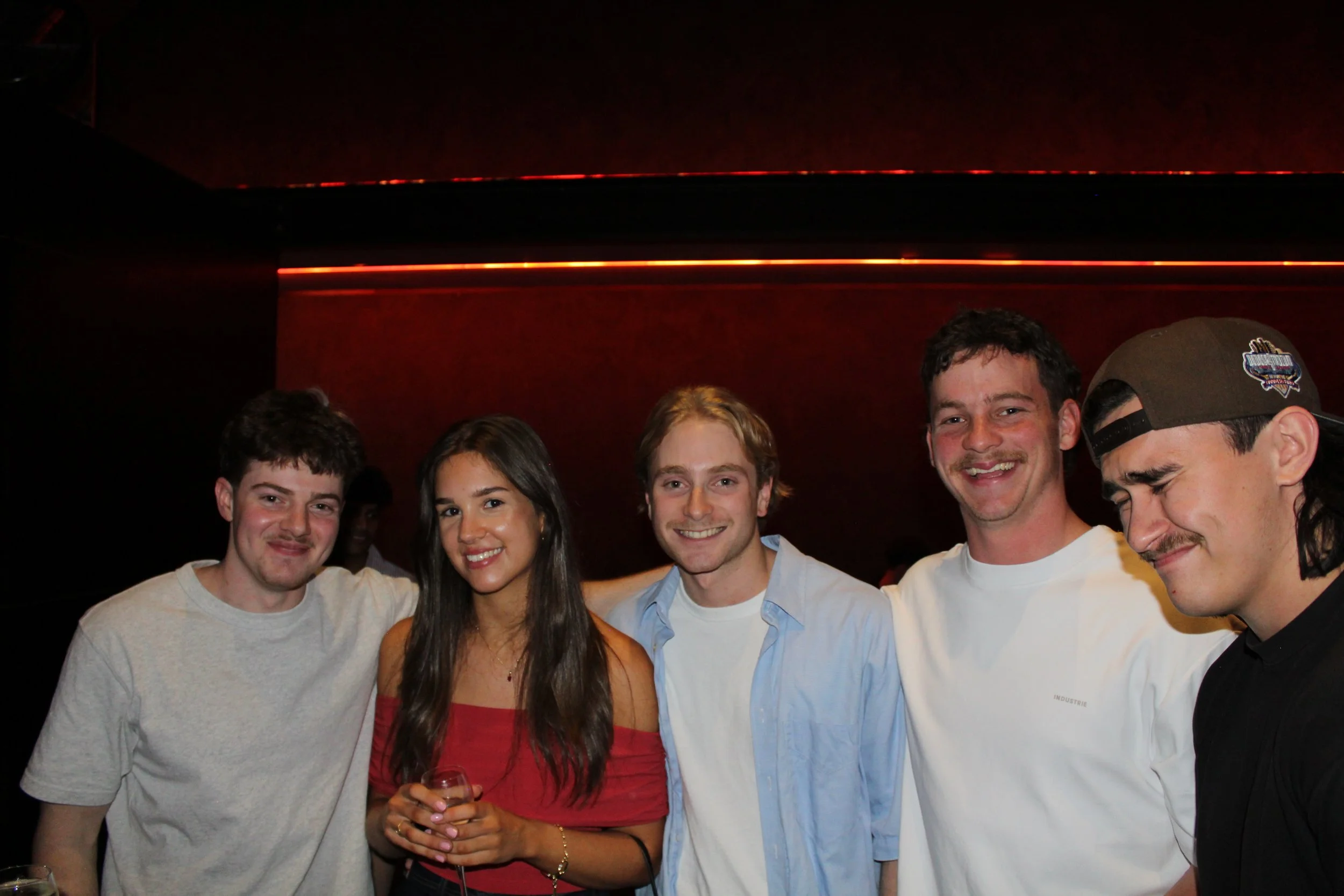 Group of five young friends smiling, posing in a dimly lit indoor setting, with a red-colored wall and ambient lighting in the background.