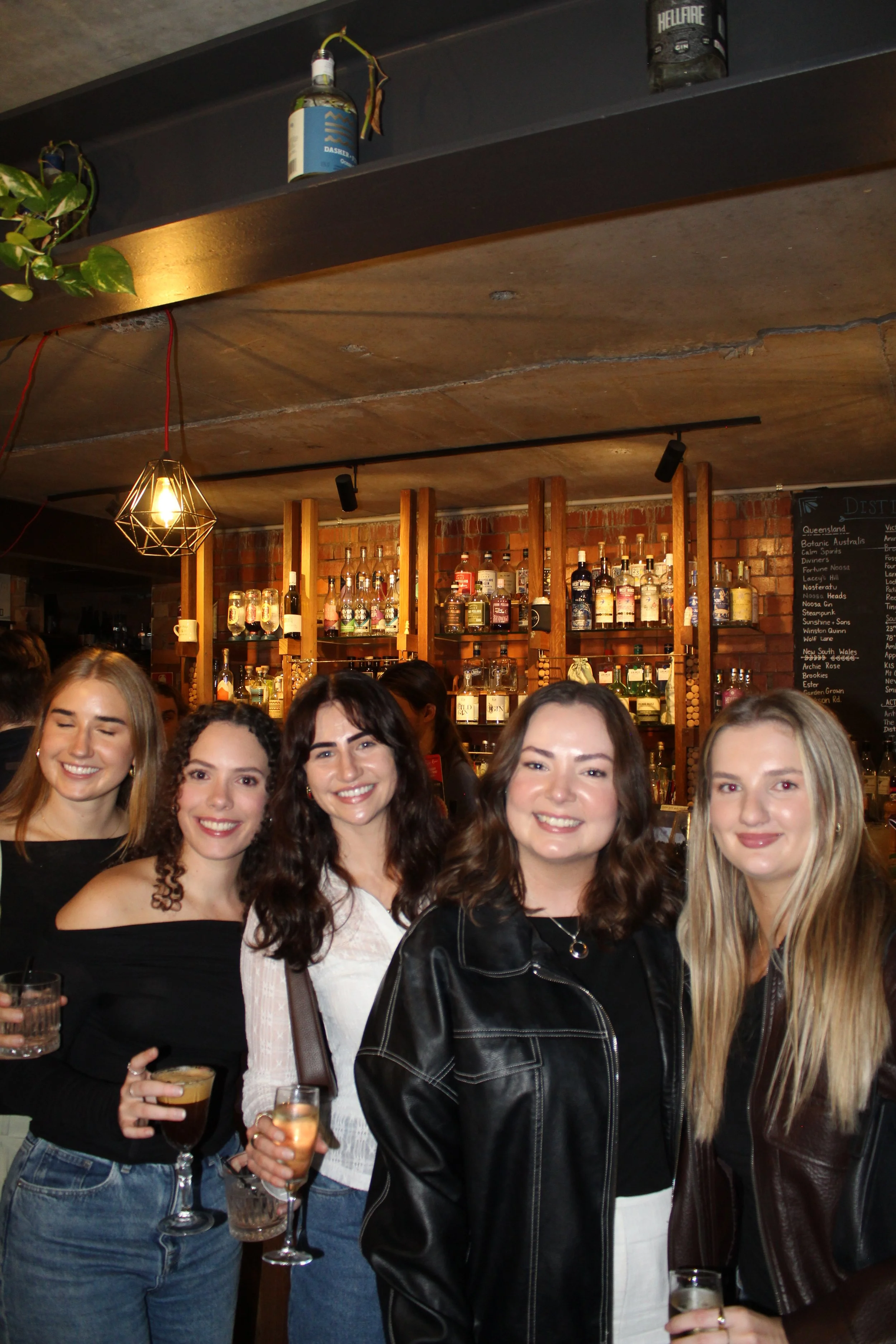 A group of women smiling and holding drinks at a bar or restaurant, with liquor bottles and a chalkboard menu in the background.