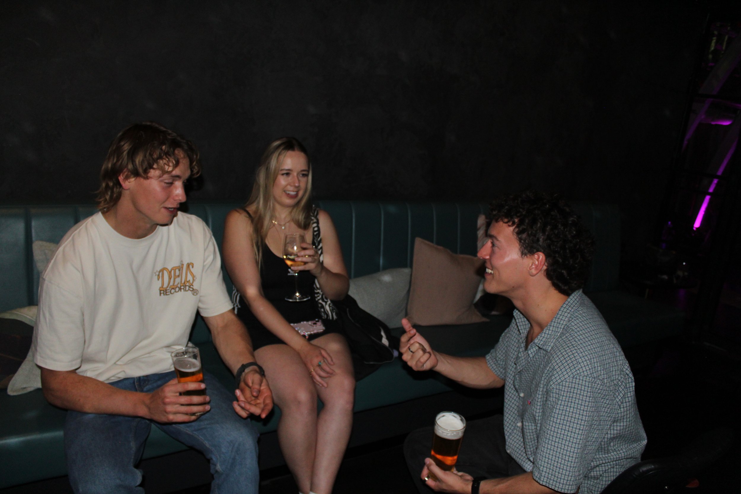 Three young adults sitting on a teal couch, drinking beers and chatting in a dimly lit bar or club with black walls.