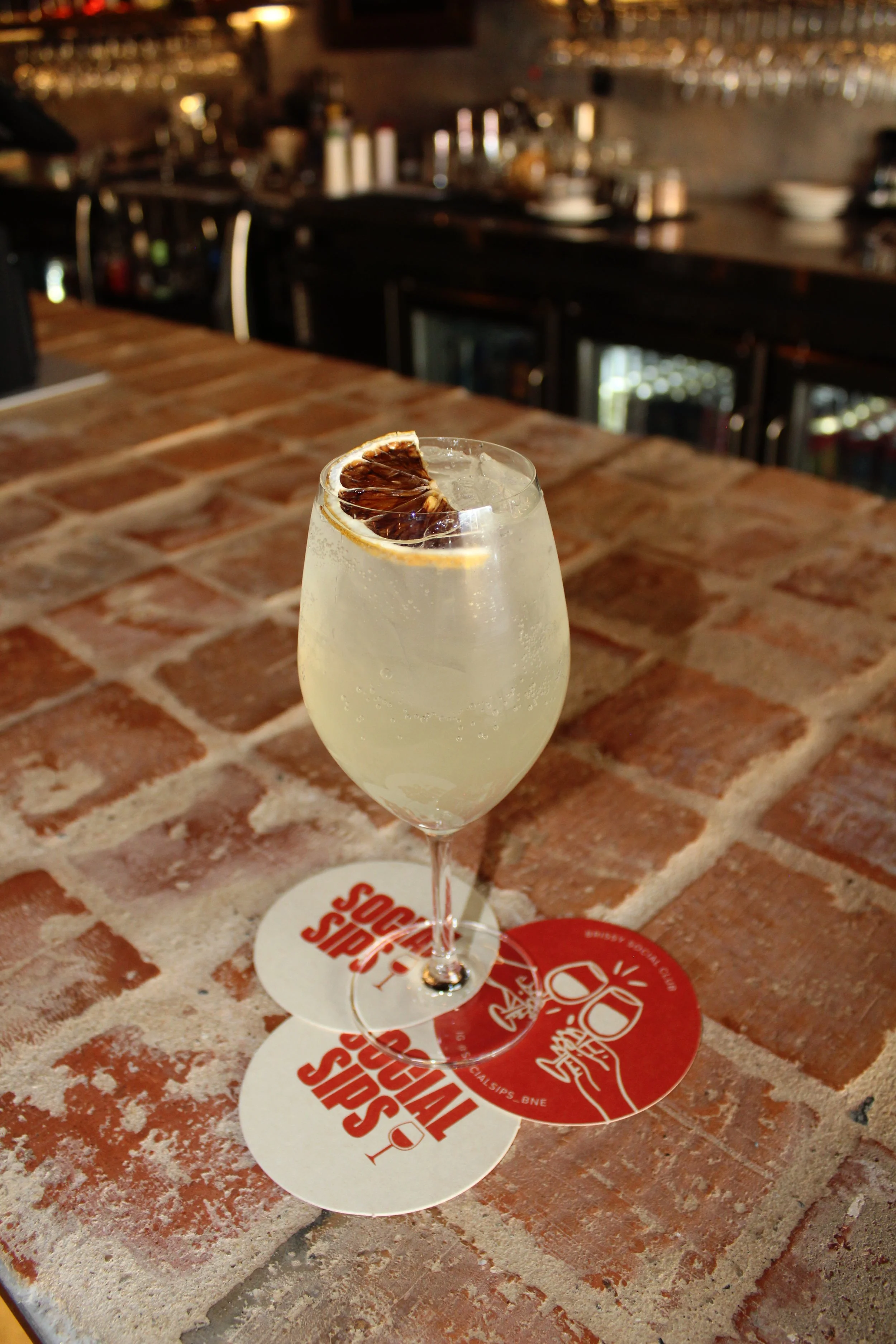 A glass of a sparkling beverage with a dried blood orange slice on top, placed on a bar countertop with two coasters underneath.