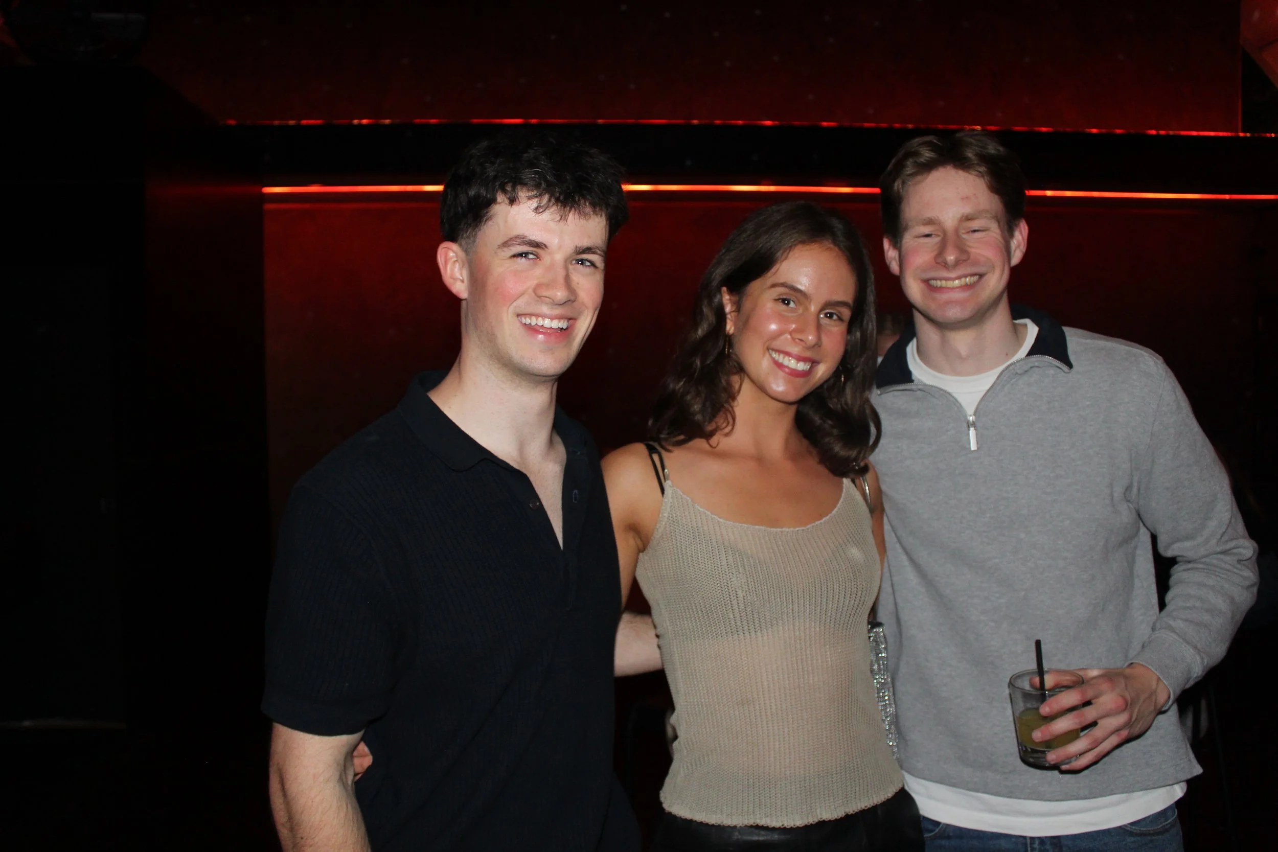 Three young adults smiling and posing together at a night event, with a dark background and red lighting.