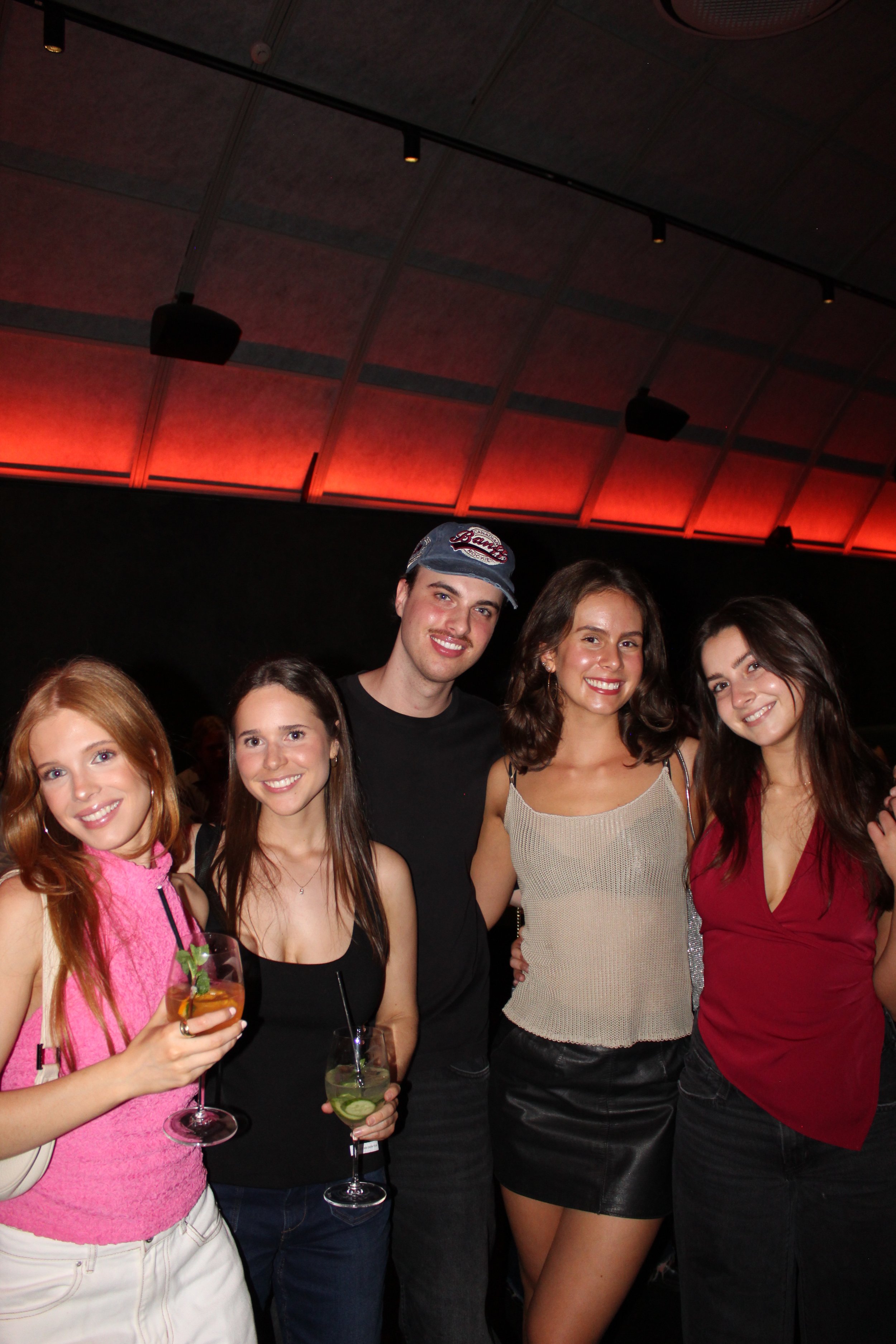 Group of five young adults posing together at a nightclub or party, holding drinks and smiling, with a dark background and red lighting overhead.