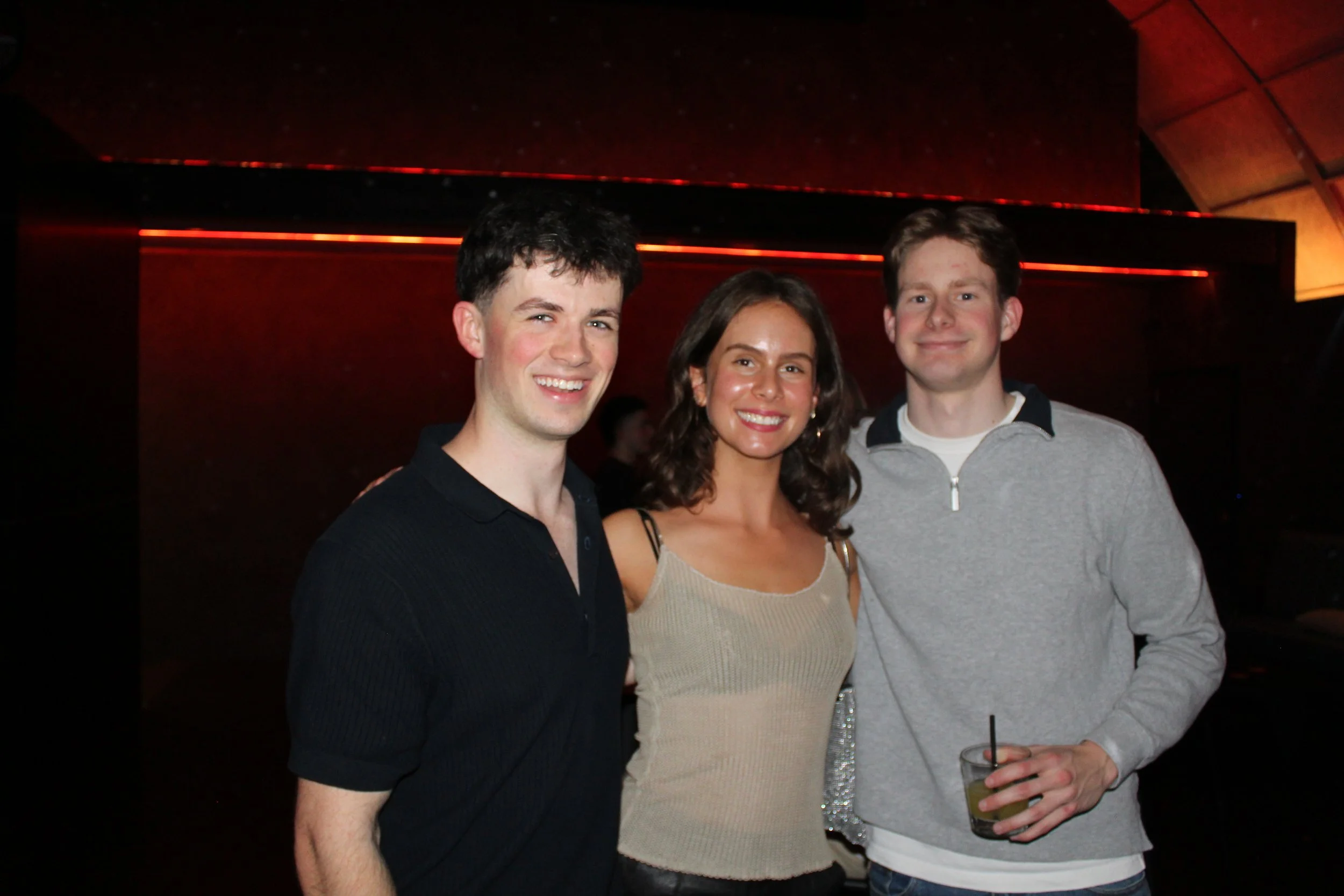 Three young adults, two men and one woman, smiling at a social gathering in a dimly lit venue. The woman is in the middle, wearing a sleeveless beige top, and the men are on either side, one in a black polo shirt and the other in a gray zip-up sweats