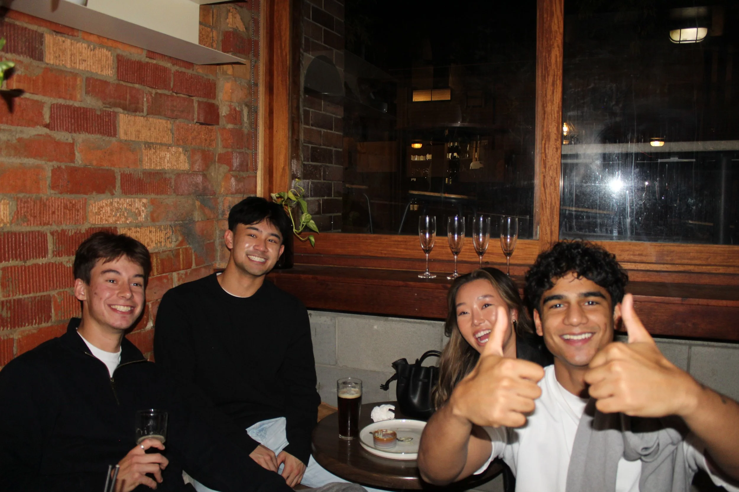 A group of smiling young people sitting together at a restaurant table, with three champagne glasses and some drinks, against a brick wall background.