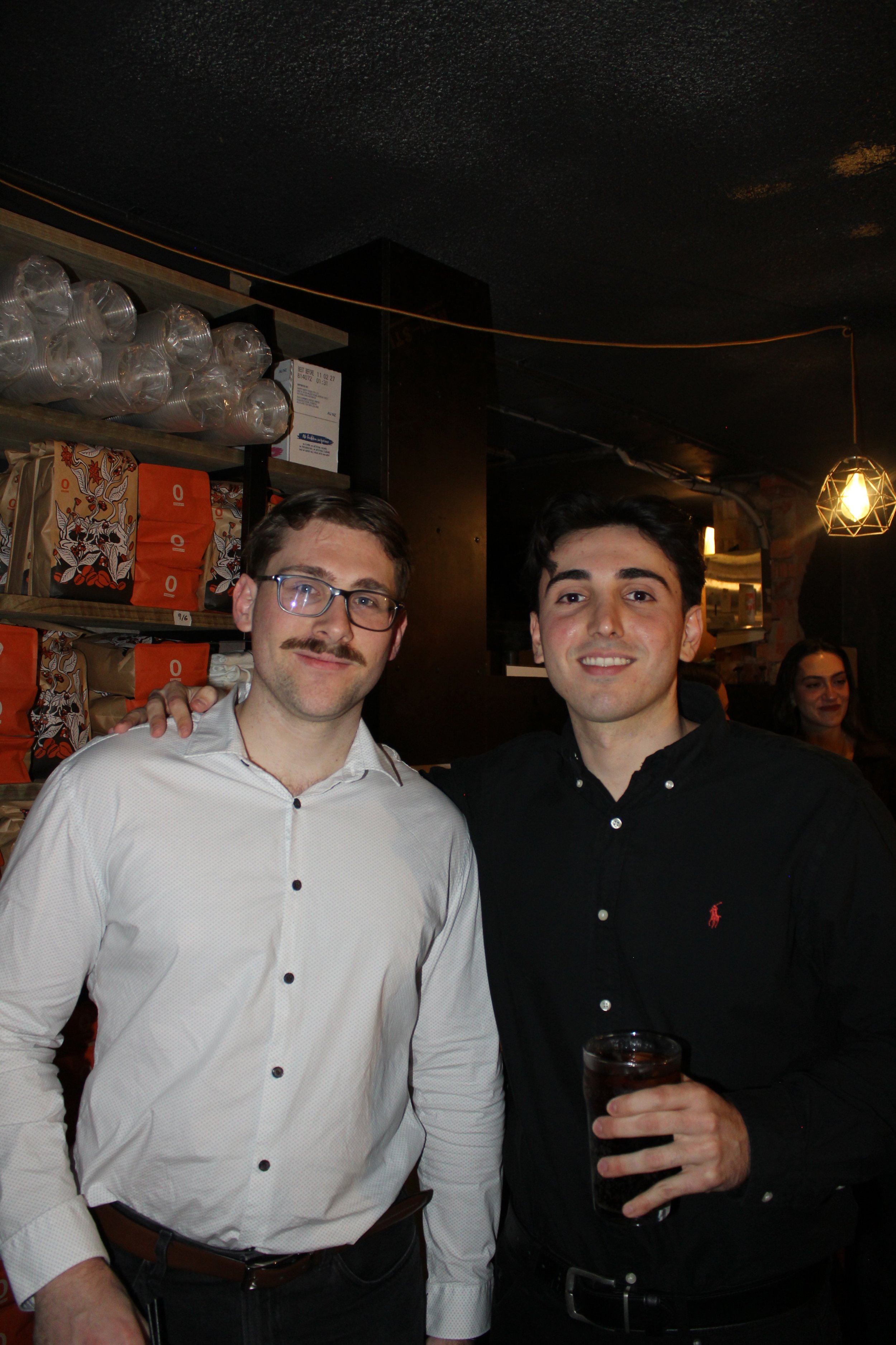 Two men standing together in a bar or restaurant, smiling at the camera. One man is wearing glasses and a white shirt, and the other is wearing a black shirt and holding a glass of dark beverage. There are shelves with items behind them and warm ligh