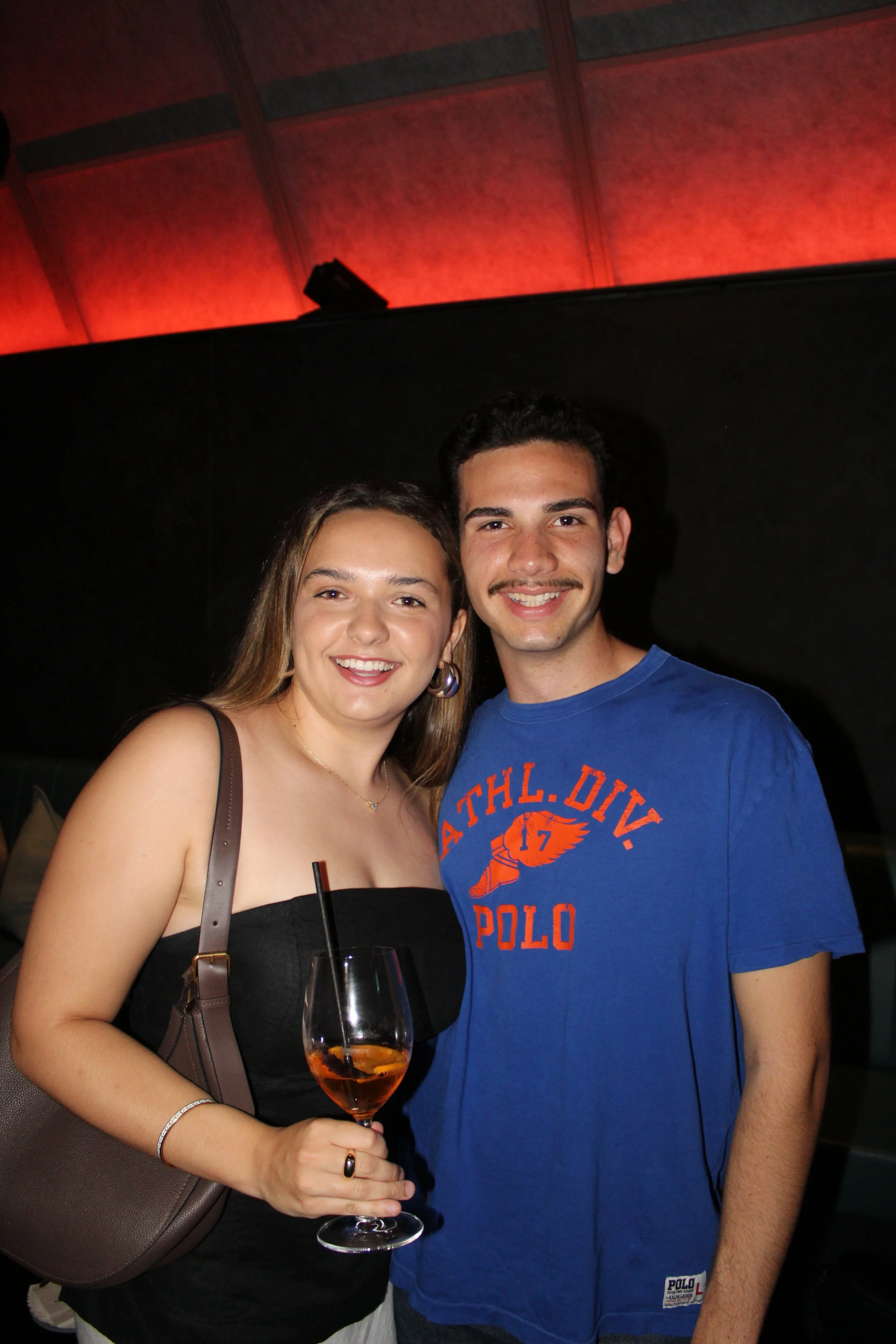 A smiling woman in a black strapless top holding a glass of orange-colored drink, standing beside a smiling man in a blue t-shirt. They are indoors with a dark background and red lighting on the ceiling.