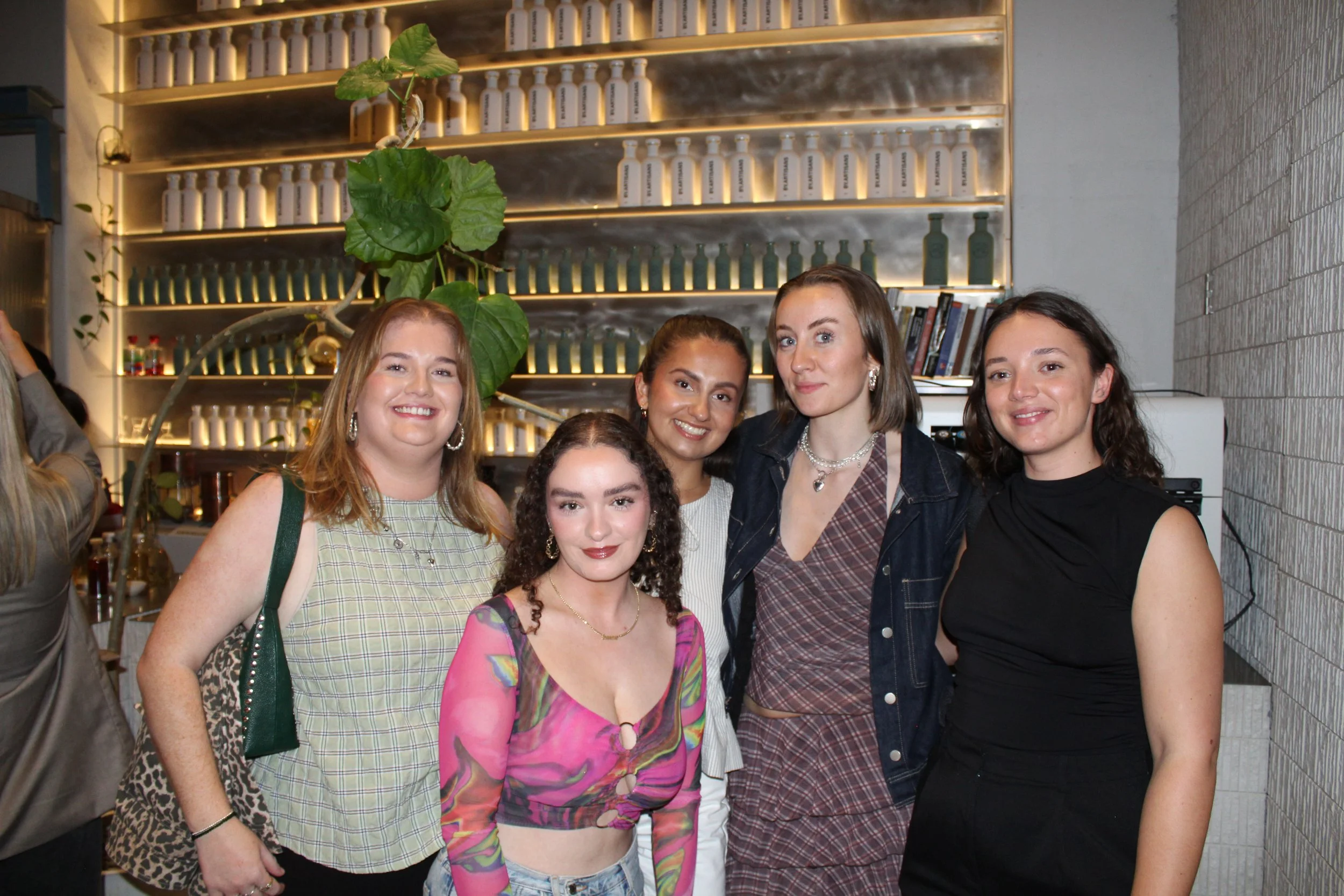 Five women posing together indoors at a social gathering, smiling at the camera with a modern decor background including shelves with bottles and books, and a large leafy plant.