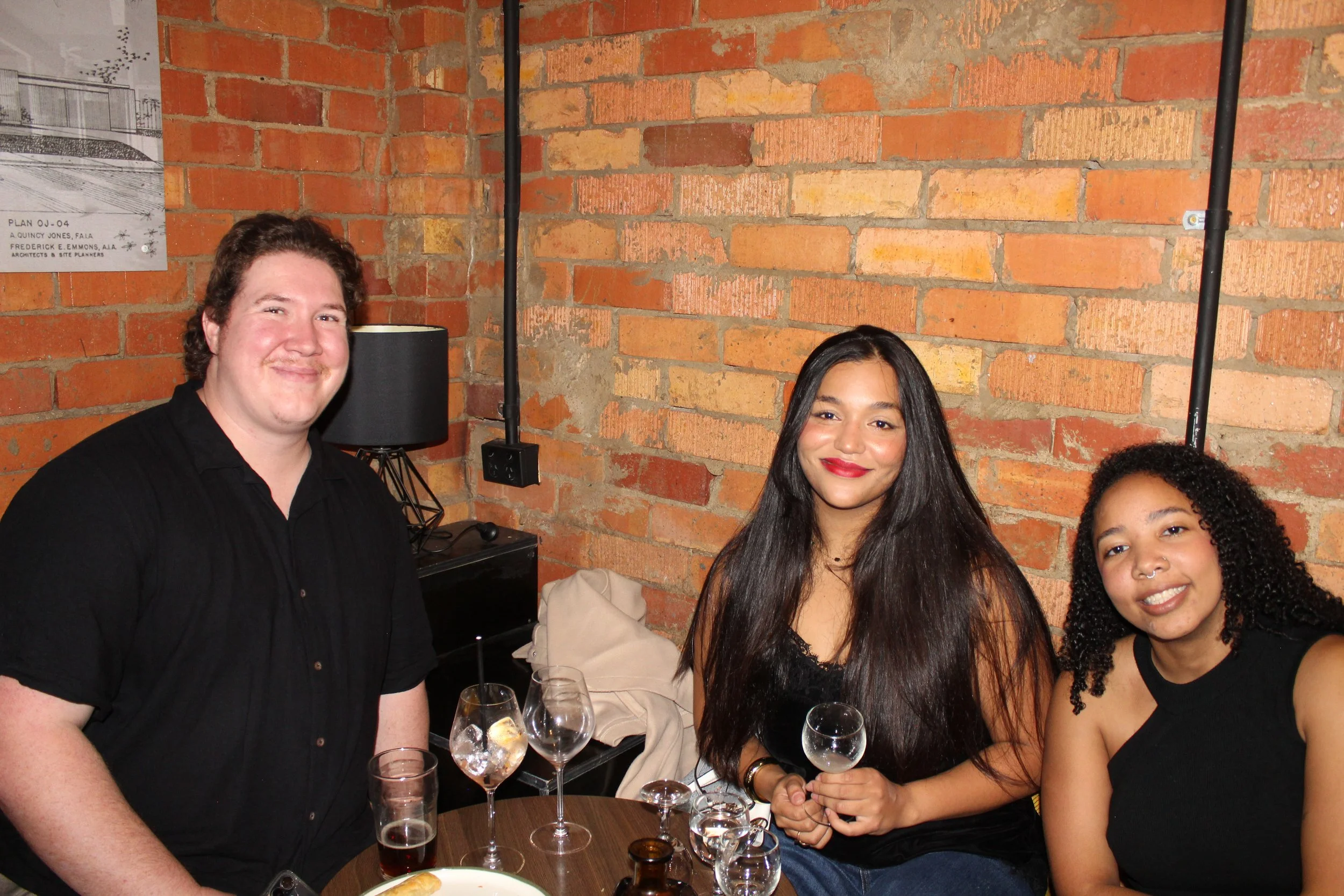 Three people gathered around a table with wine glasses, in front of a brick wall, in a dimly lit room, smiling for the camera.