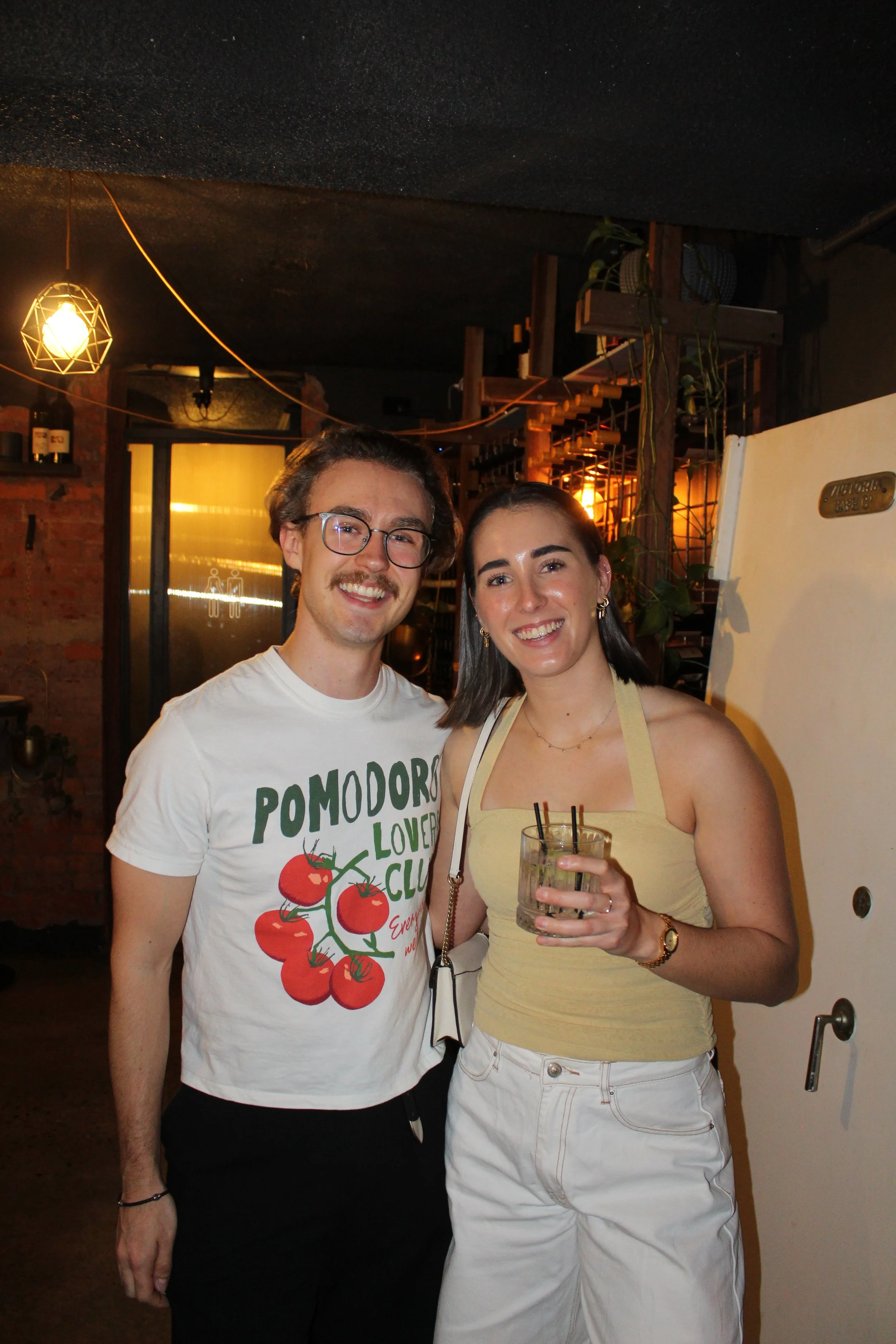 A young man with glasses and a young woman holding a drink standing in a dimly lit bar or restaurant with warm lighting, brick wall, and wooden decor.