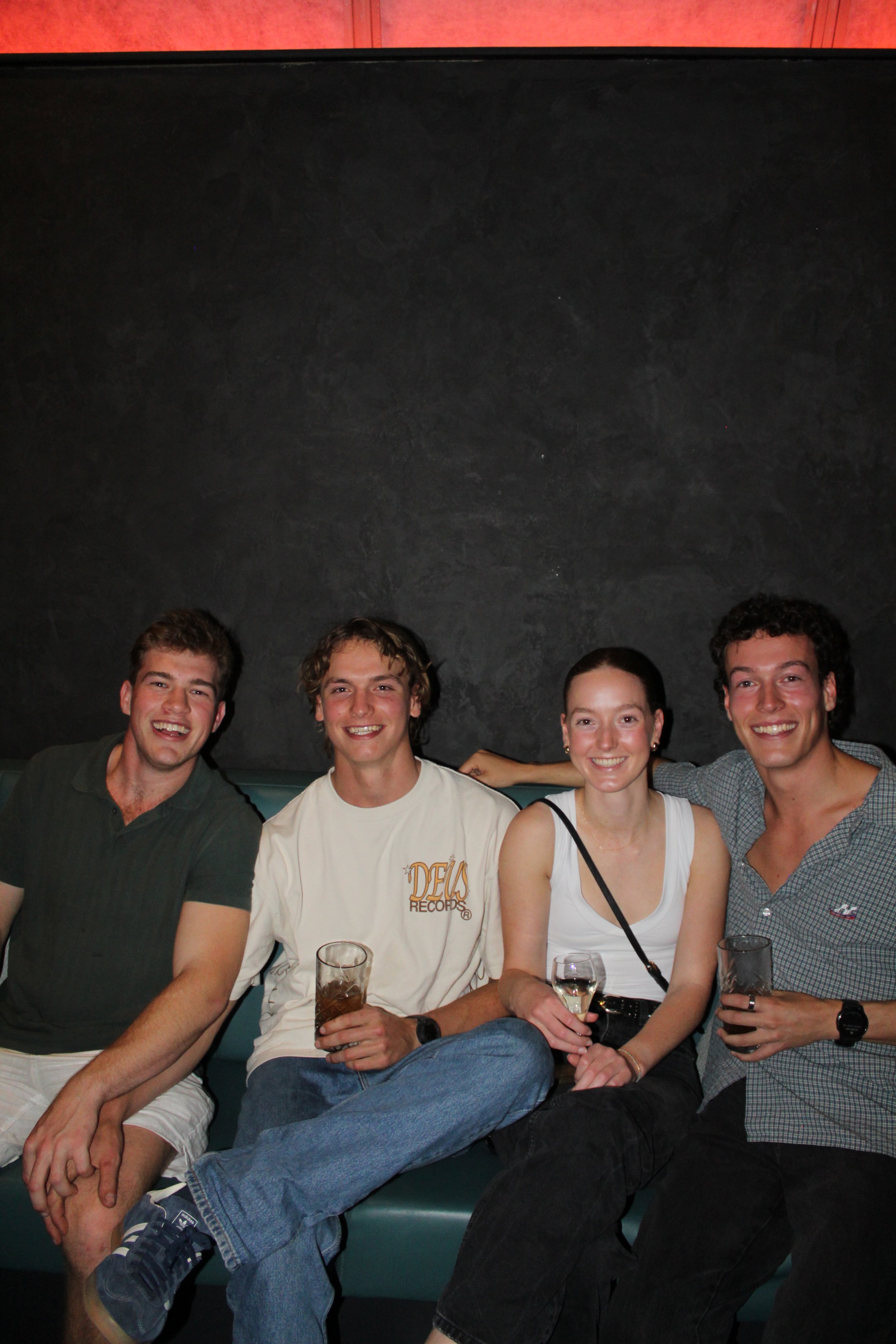 Four young adults sitting on a teal-colored bench in front of a dark wall, smiling, holding drinks, and enjoying a night out.