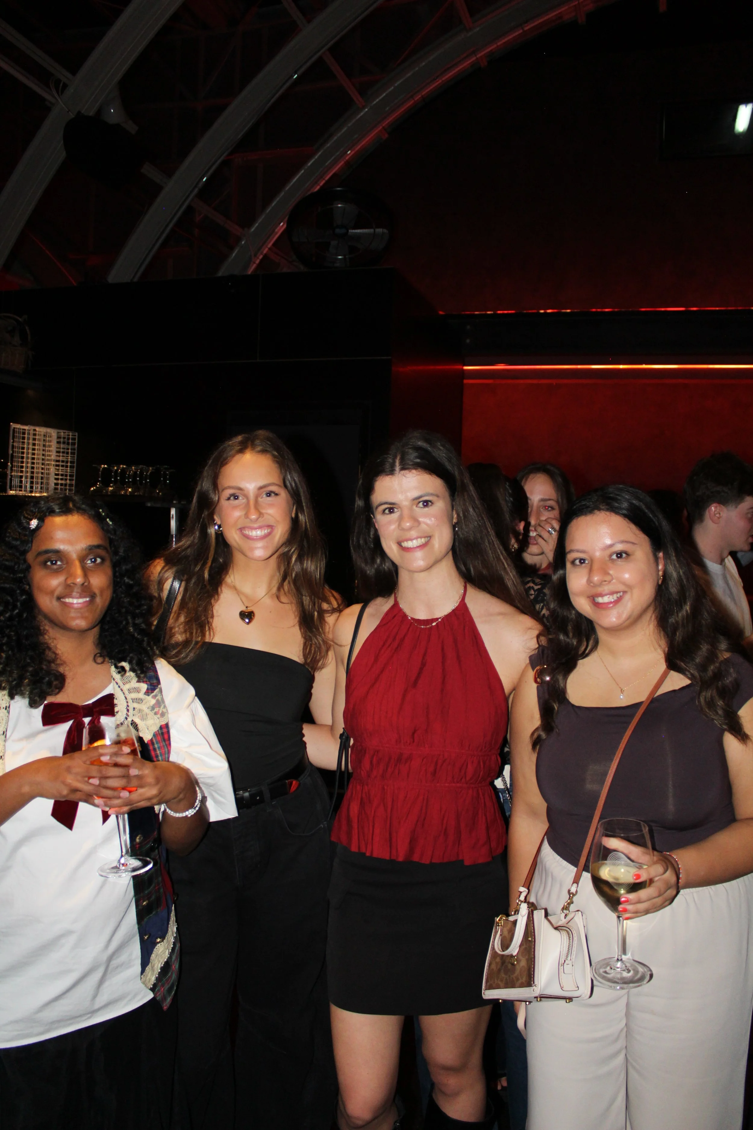 Group of four women smiling at a social event, holding drinks inside a dimly lit venue with modern decor.