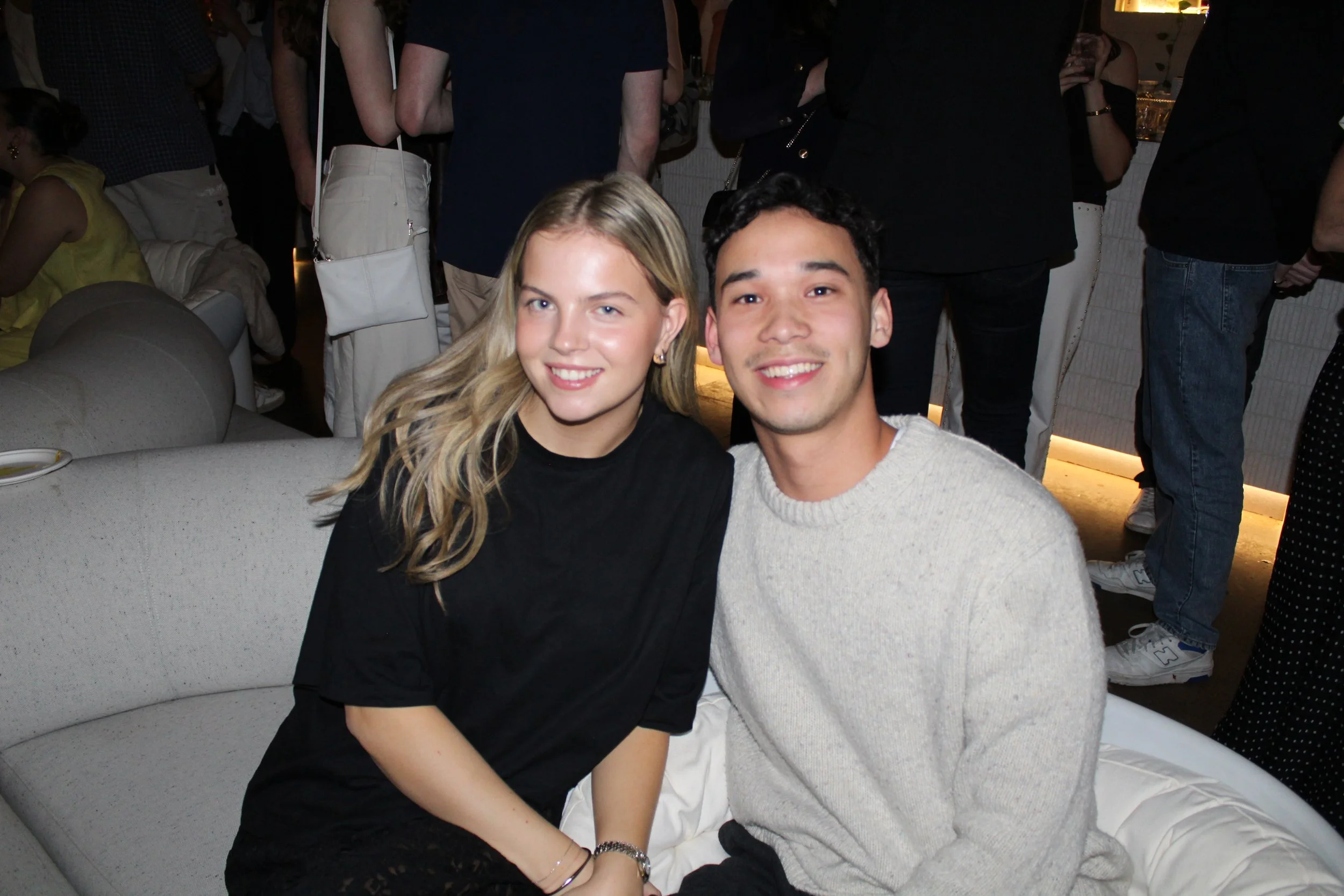 A young woman with long blonde hair and a young man with short dark hair sitting close together, smiling at the camera, at a crowded social event.