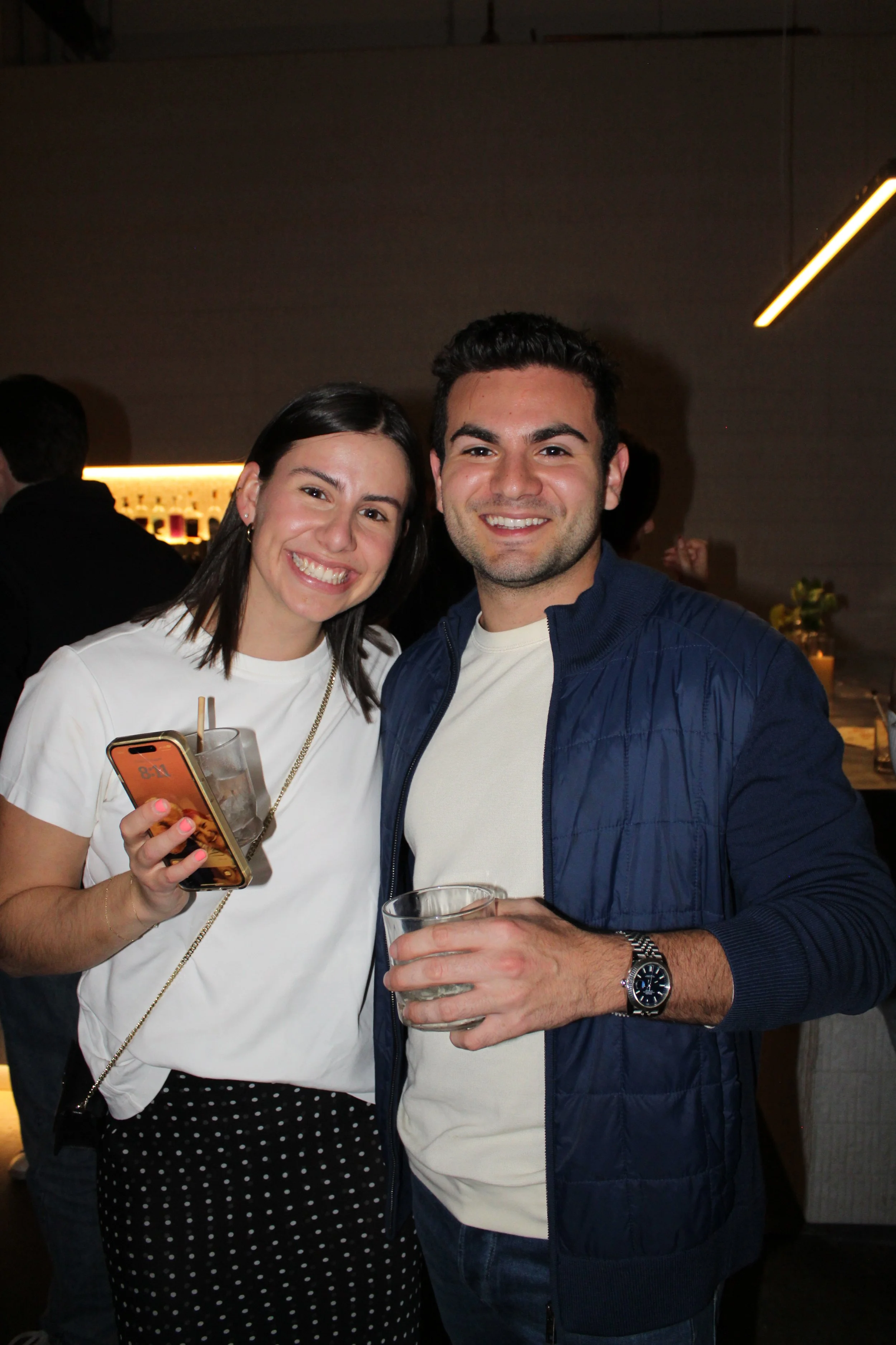 A happy woman and man smiling at the camera at a social gathering, holding drinks in a dimly lit setting.