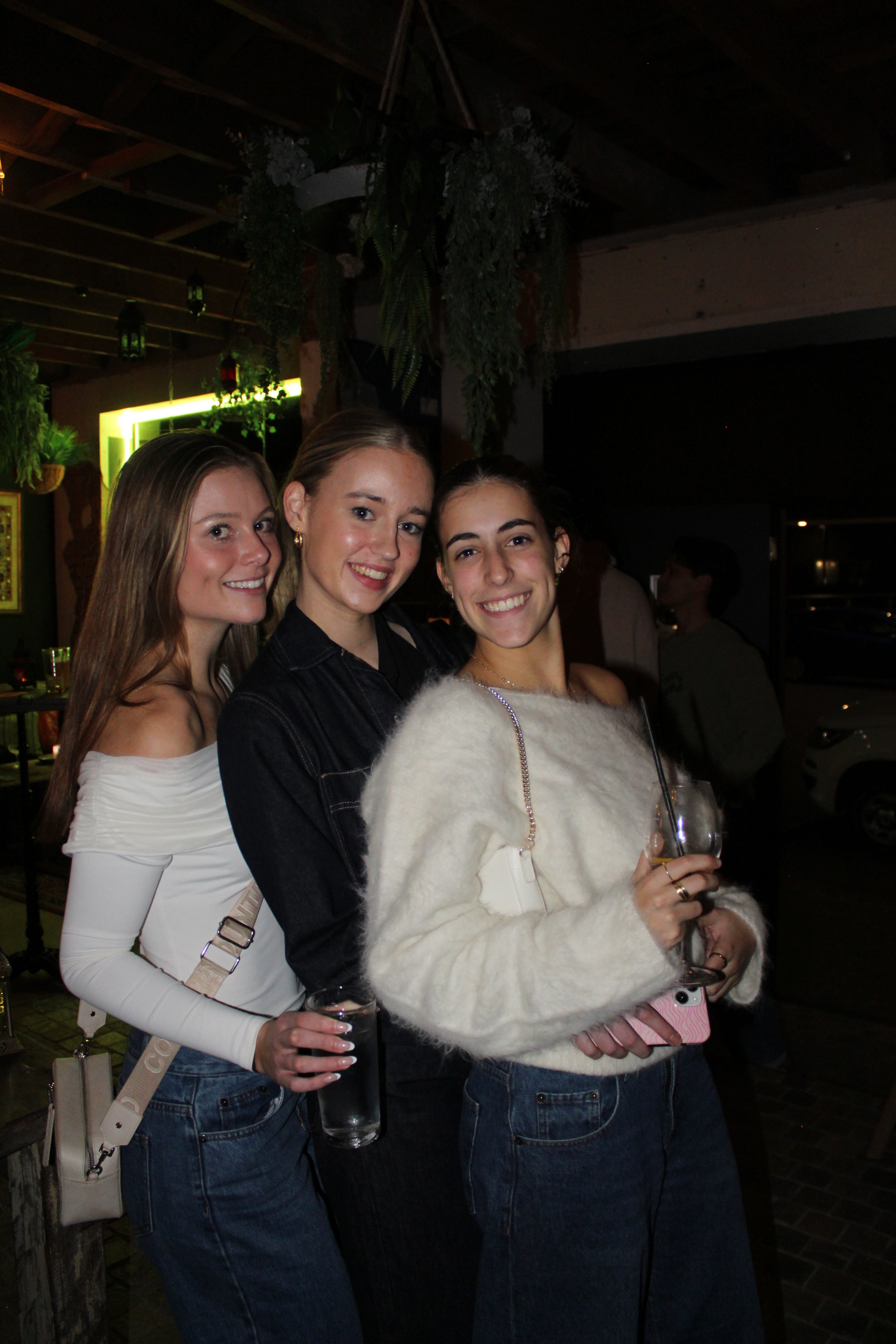 Three young women smiling and holding drinks at a party or gathering indoors at night.
