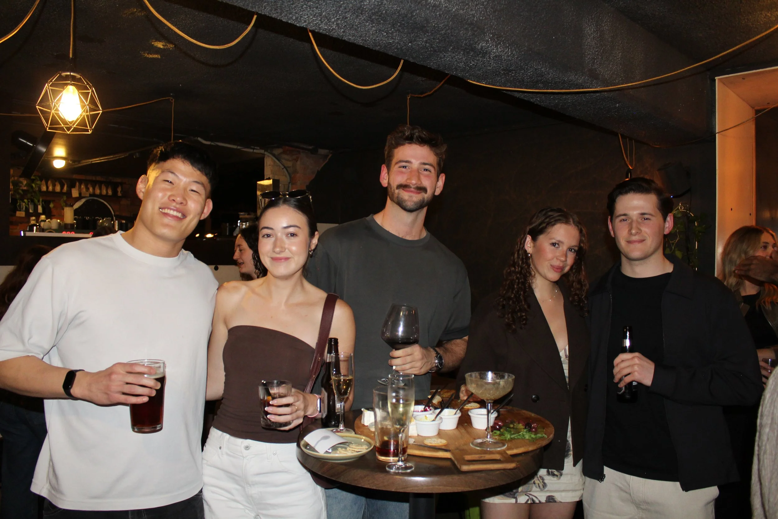 A group of six young adults posing at a social gathering in a dimly lit indoor venue. They are smiling, holding drinks, and standing behind a table with food and drinks.