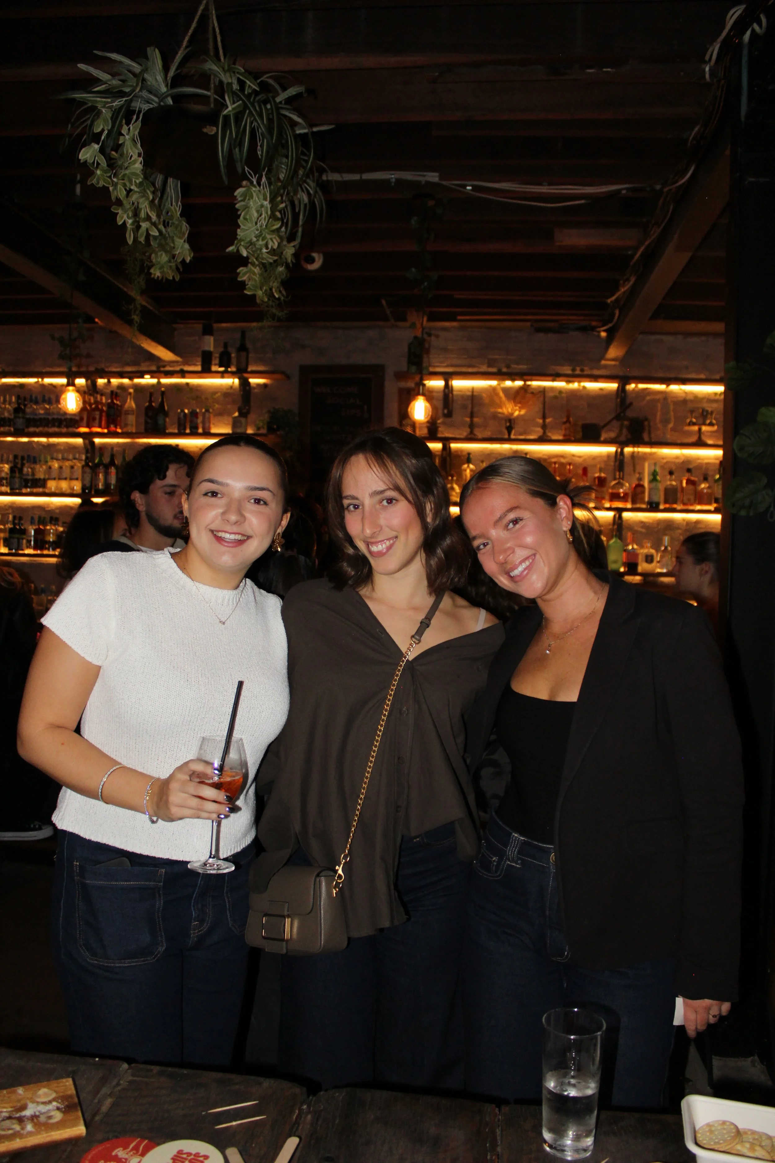 Three women smiling at a bar or restaurant, with shelves of liquor bottles and warm lighting in the background.