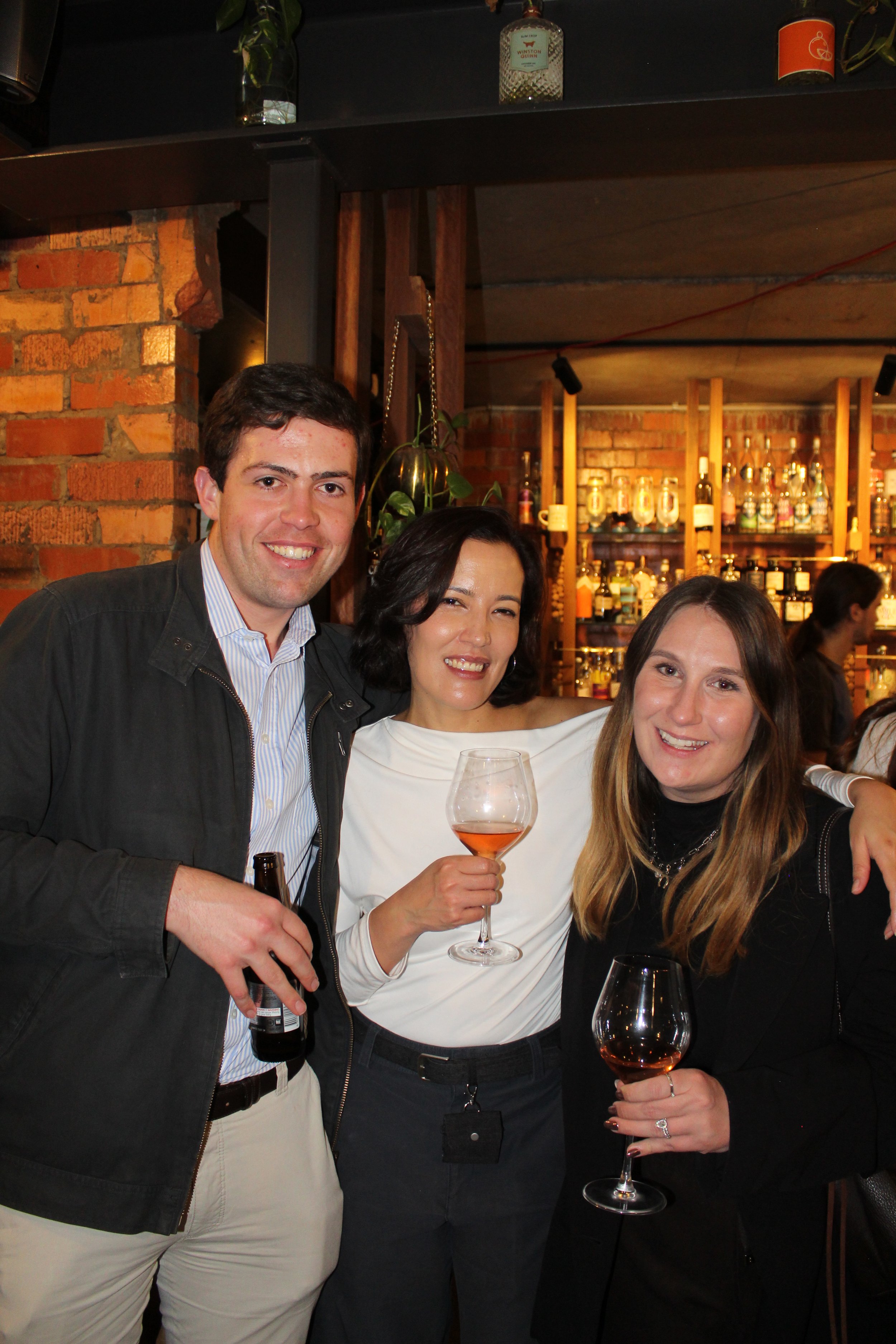 Three people smiling at a bar, holding drinks, with a background of shelves filled with liquor bottles.