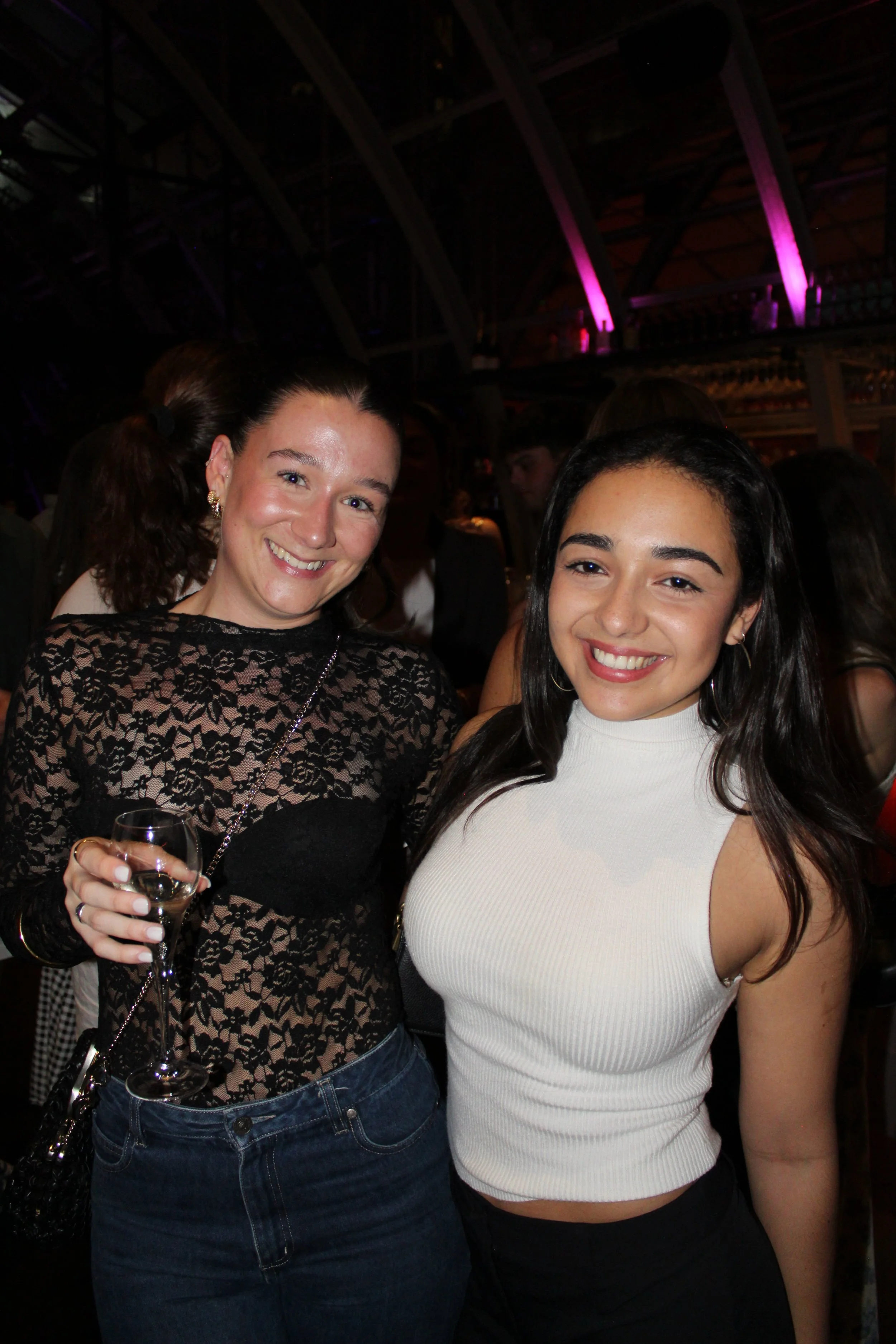 Two women smiling at a social event, one holding a glass of wine.
