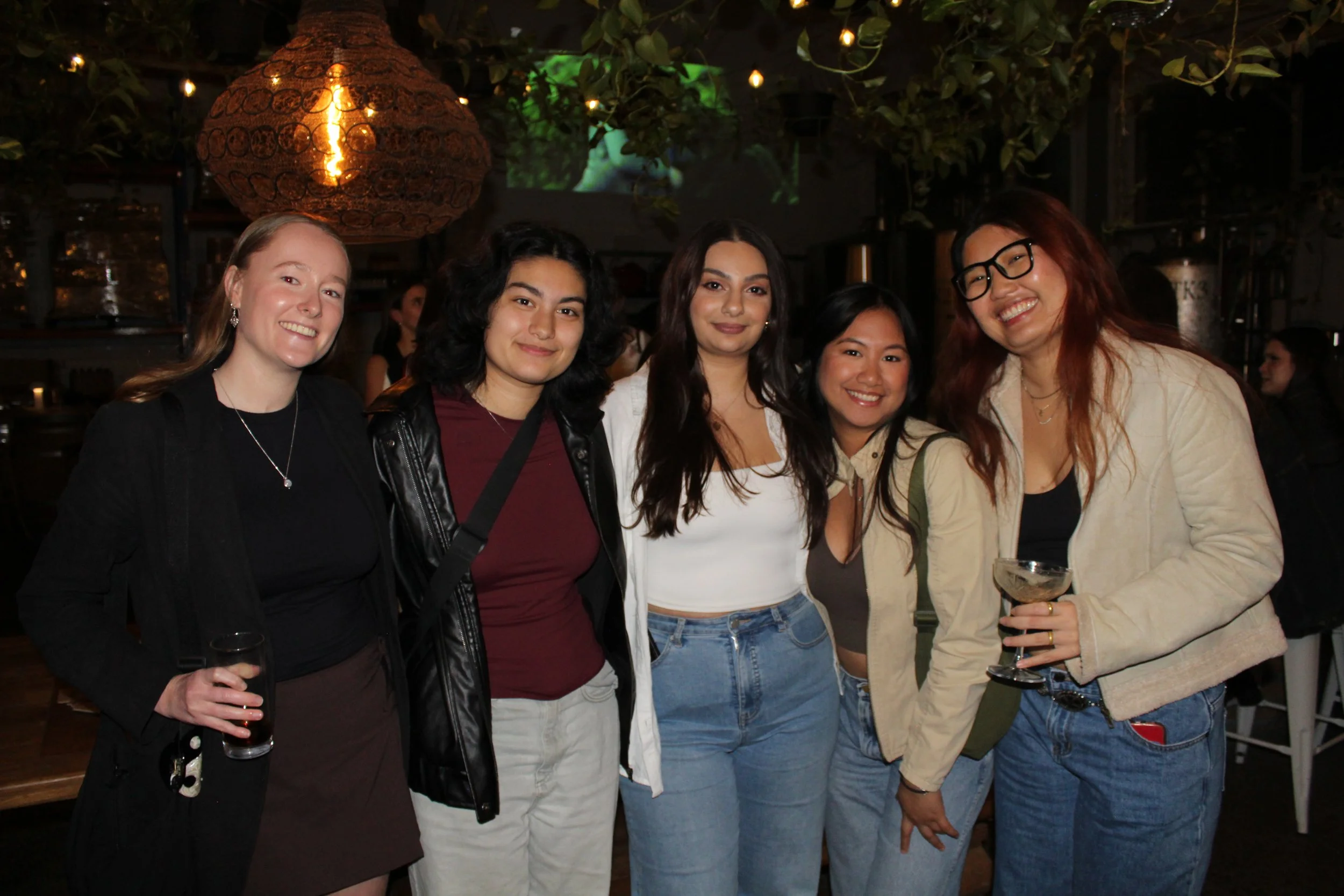 A group of five women smiling and posing together at a social gathering in a dimly lit venue, with some holding drinks.