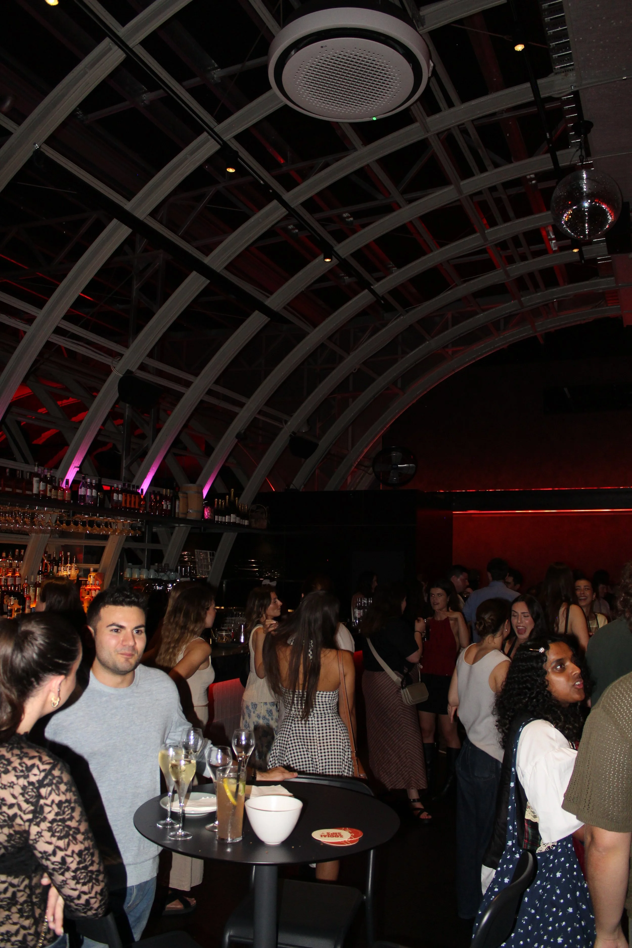 People socializing and dancing in a nightclub with a bar, curved ceiling structure, and disco ball.