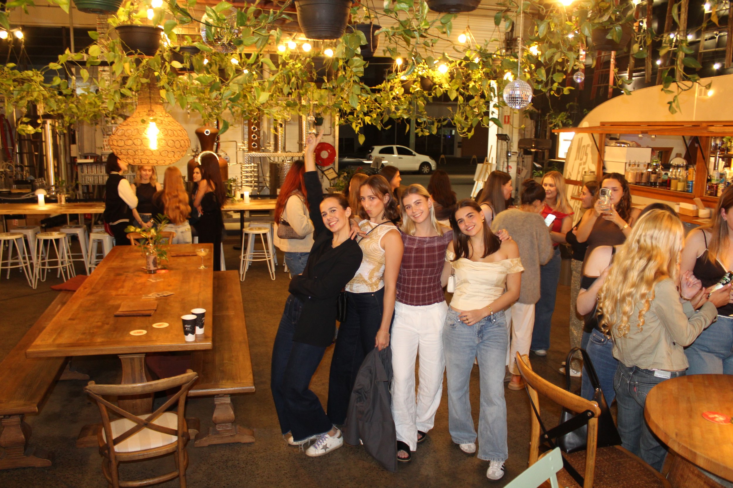 A group of five young women standing together and smiling for a photo in a lively indoor bar or restaurant with wooden furniture, hanging lights, and greenery ceiling decor. Other patrons are visible in the background.
