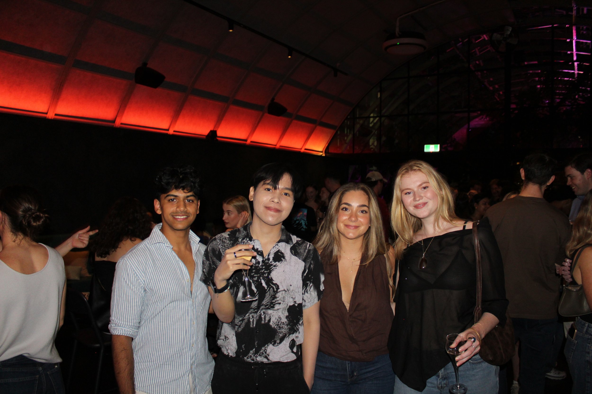 Four friends at a party, smiling and holding drinks, with a dark, colorful-lit venue in the background.