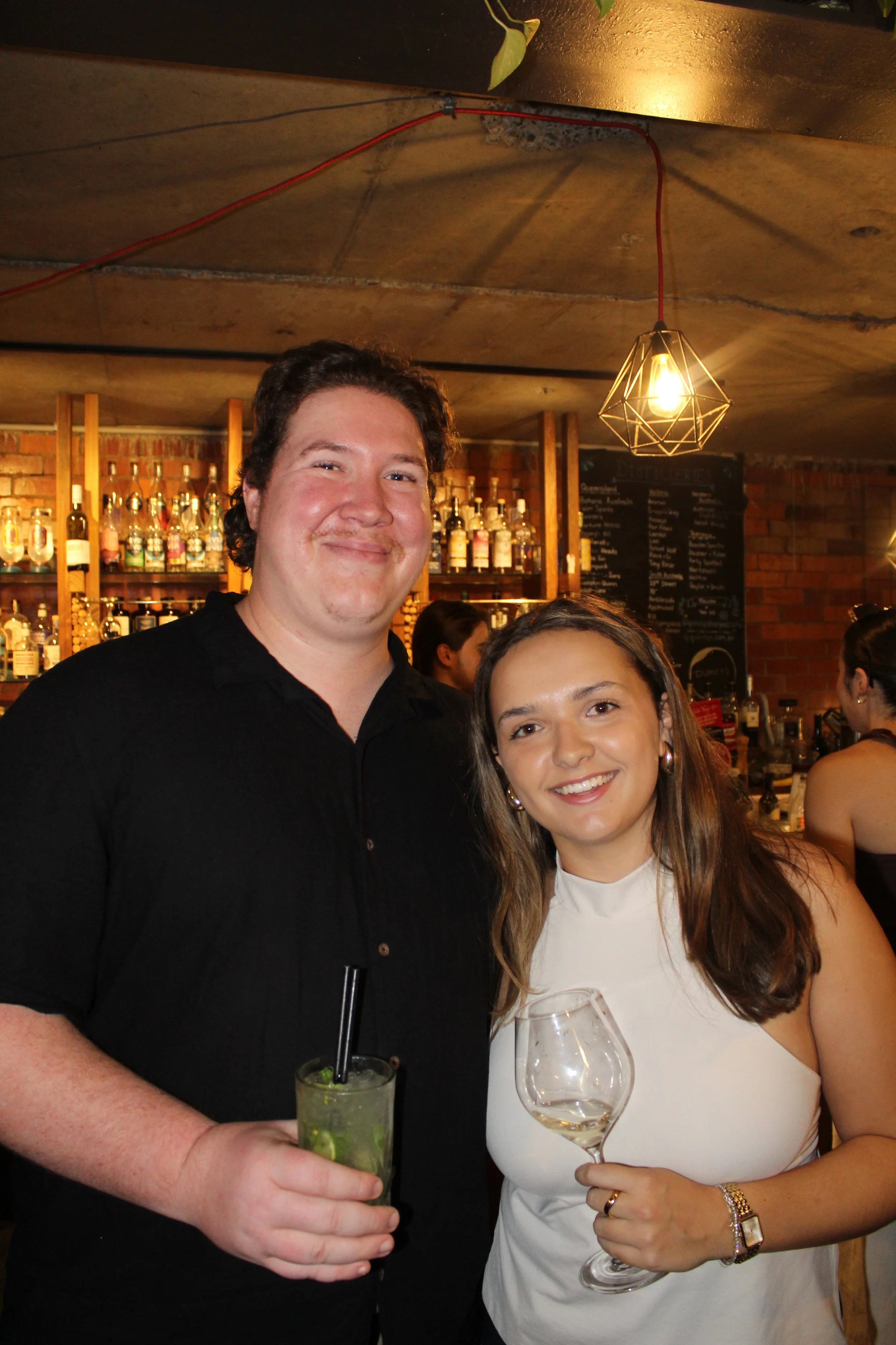 Smiling man and woman holding drinks in a restaurant with a bar and wine menu in the background.
