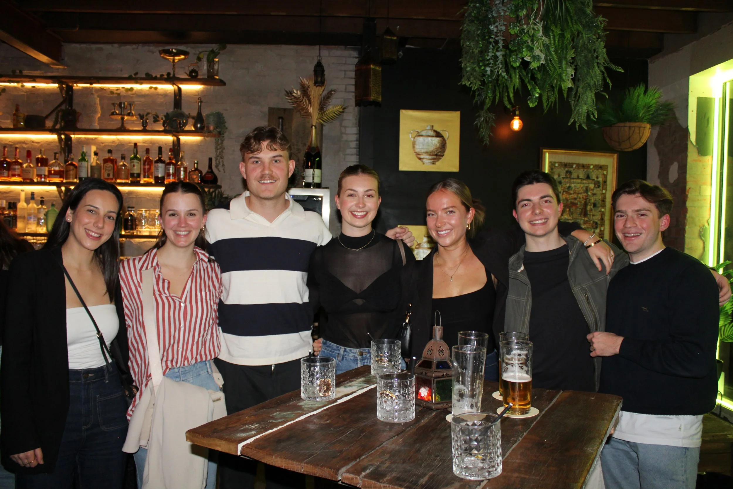 Group of seven young adults smiling, standing around a wooden table in a bar or pub with drinks, shelves with bottles and decorative plants in the background.