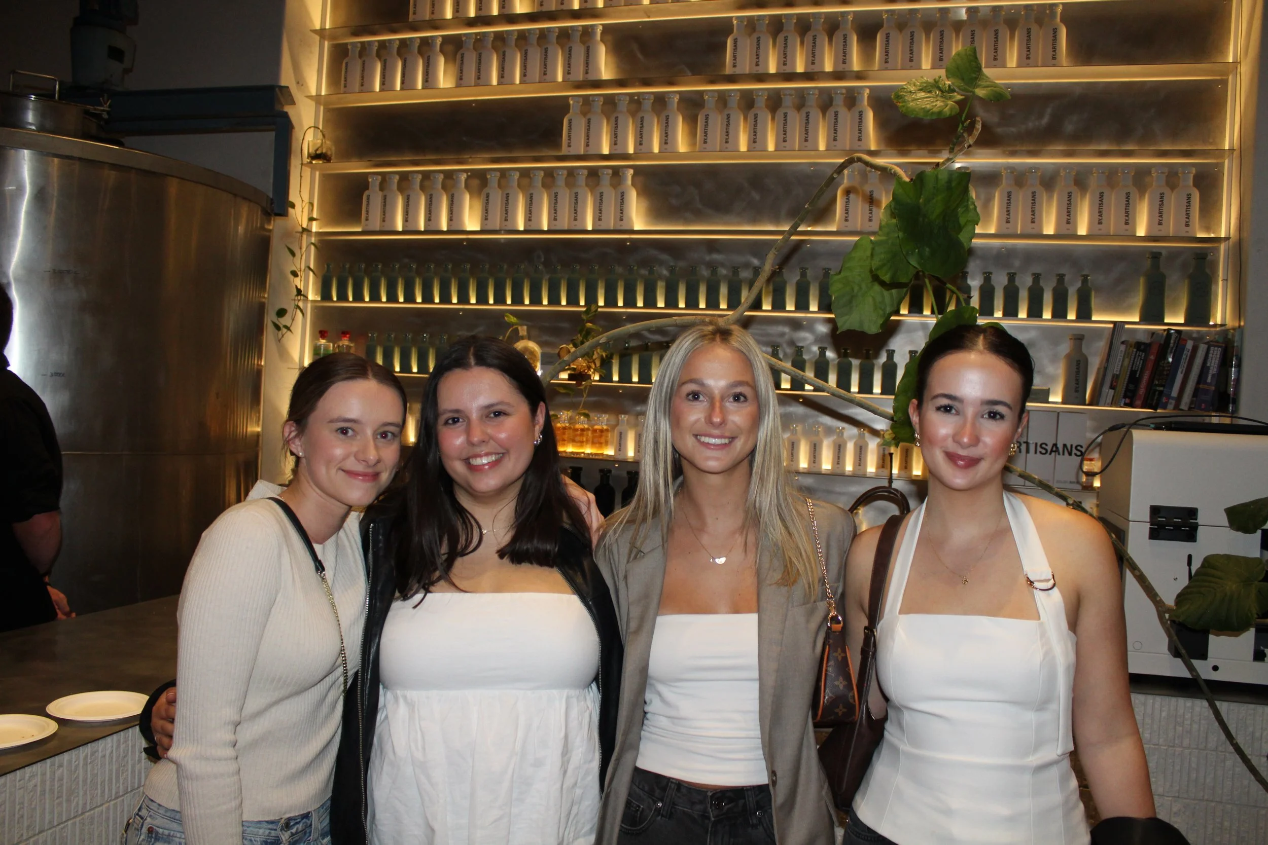 Four women standing in a cafe or restaurant, smiling at the camera, with shelves of bottles and books behind them and a large plant.