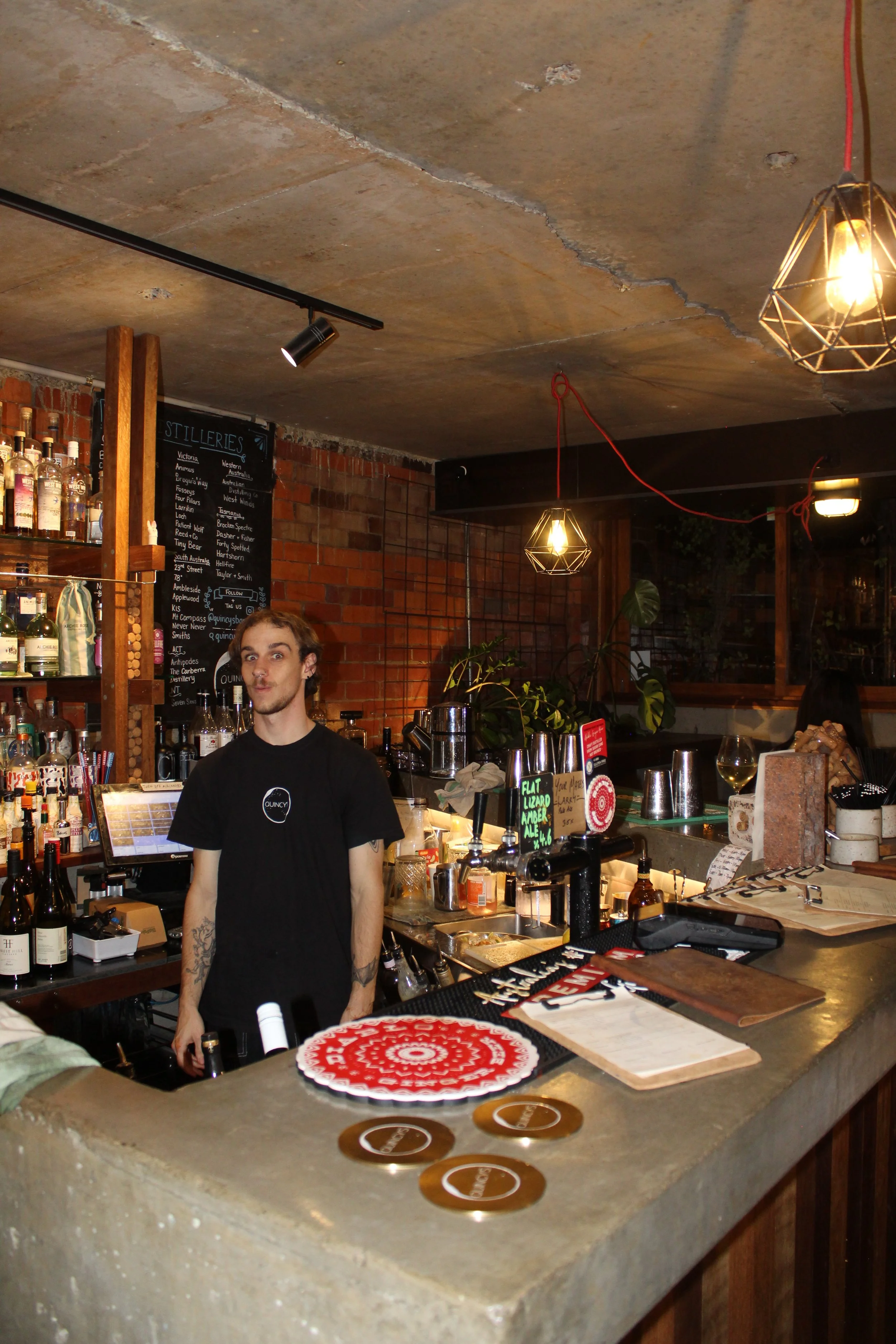 A man with tattoos wearing a black t-shirt standing behind a bar counter in a restaurant or bar. The bar counter has various bottles, a menu, and decorative coasters. The background features a brick wall with a black chalkboard menu listing various w