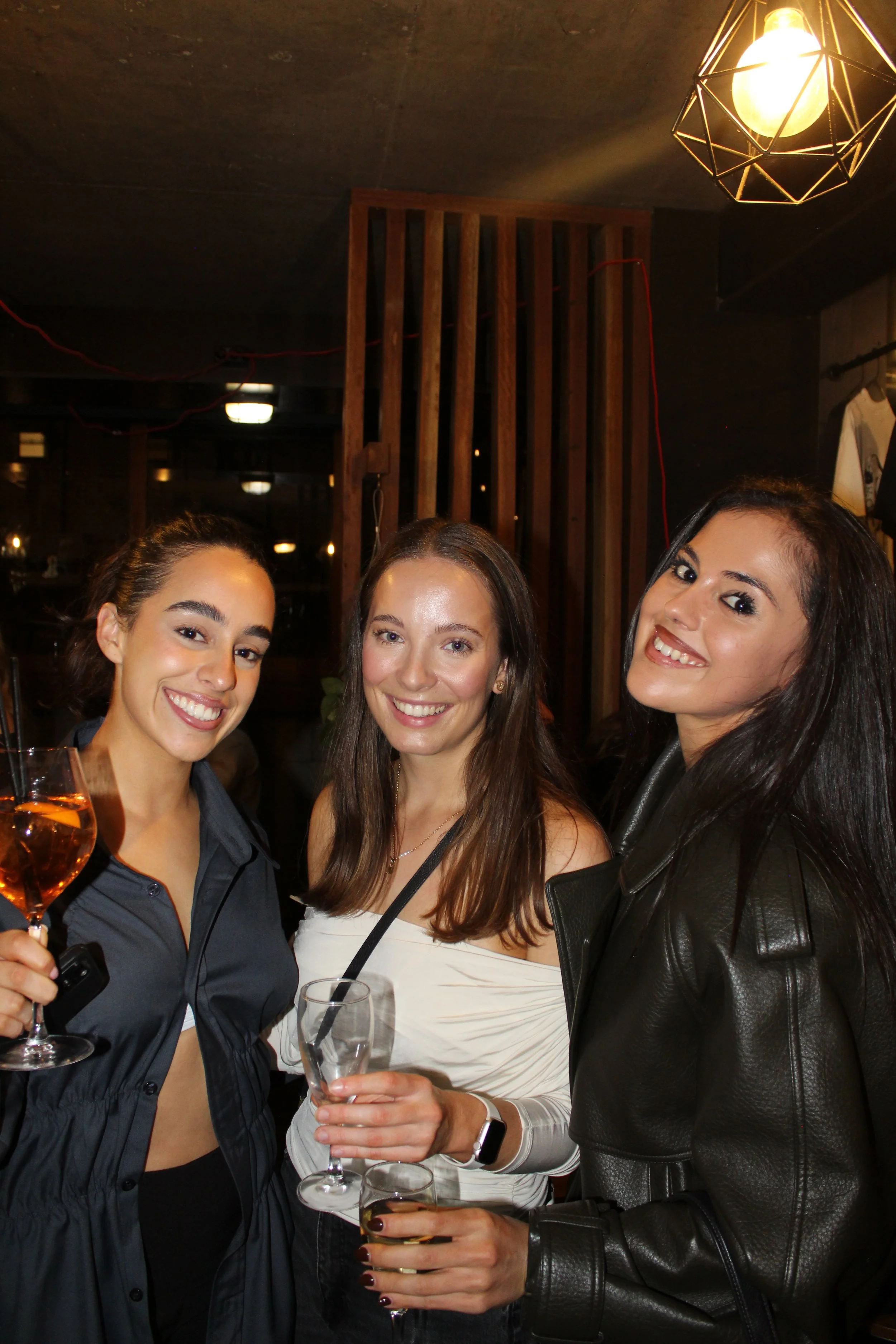 Three women smiling at a social gathering, holding wine glasses. The background features wooden decor, warm lighting, and a hanging light fixture.