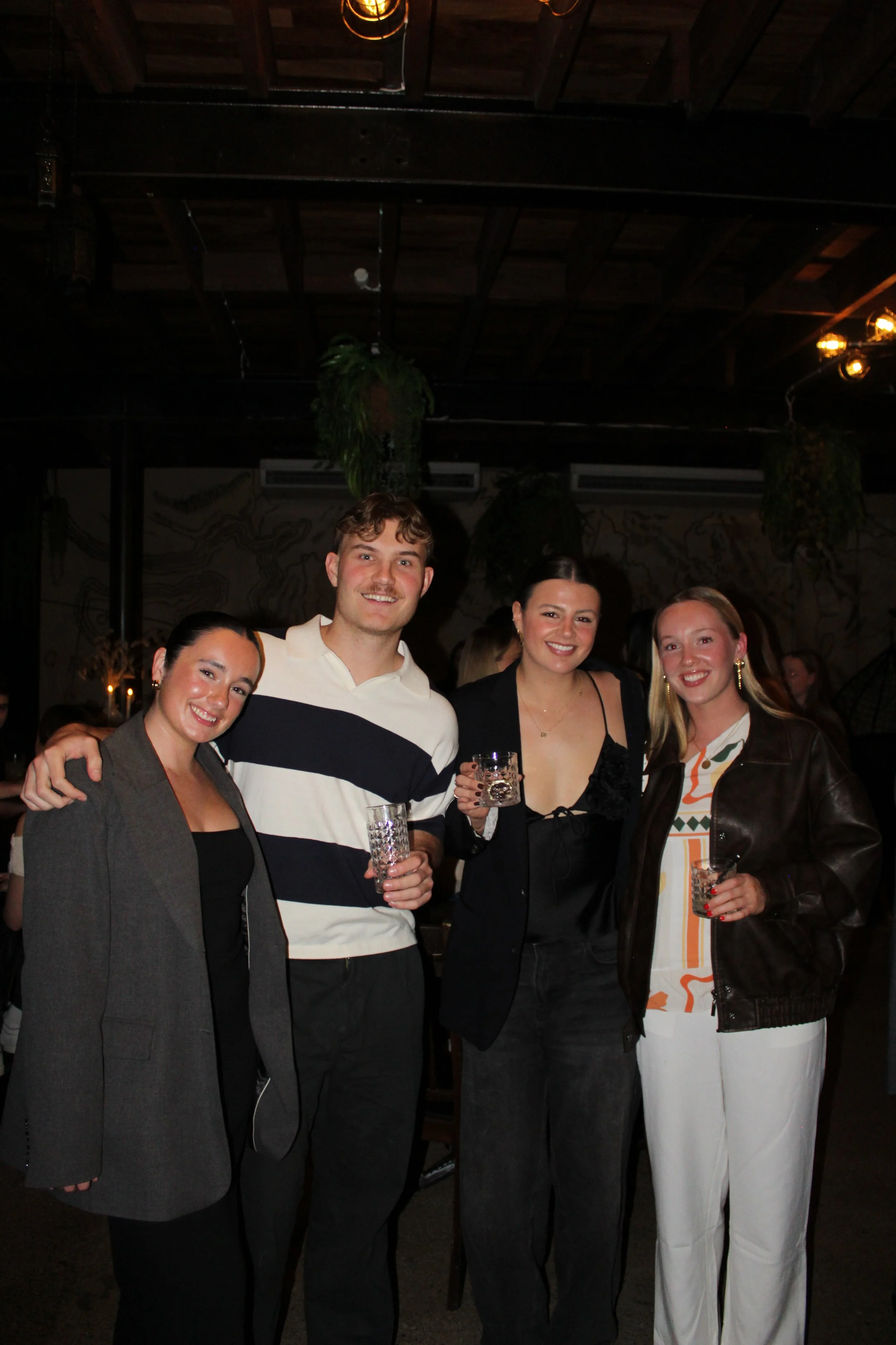 Four people smiling and holding drinks at a social event in a dimly lit indoor venue.