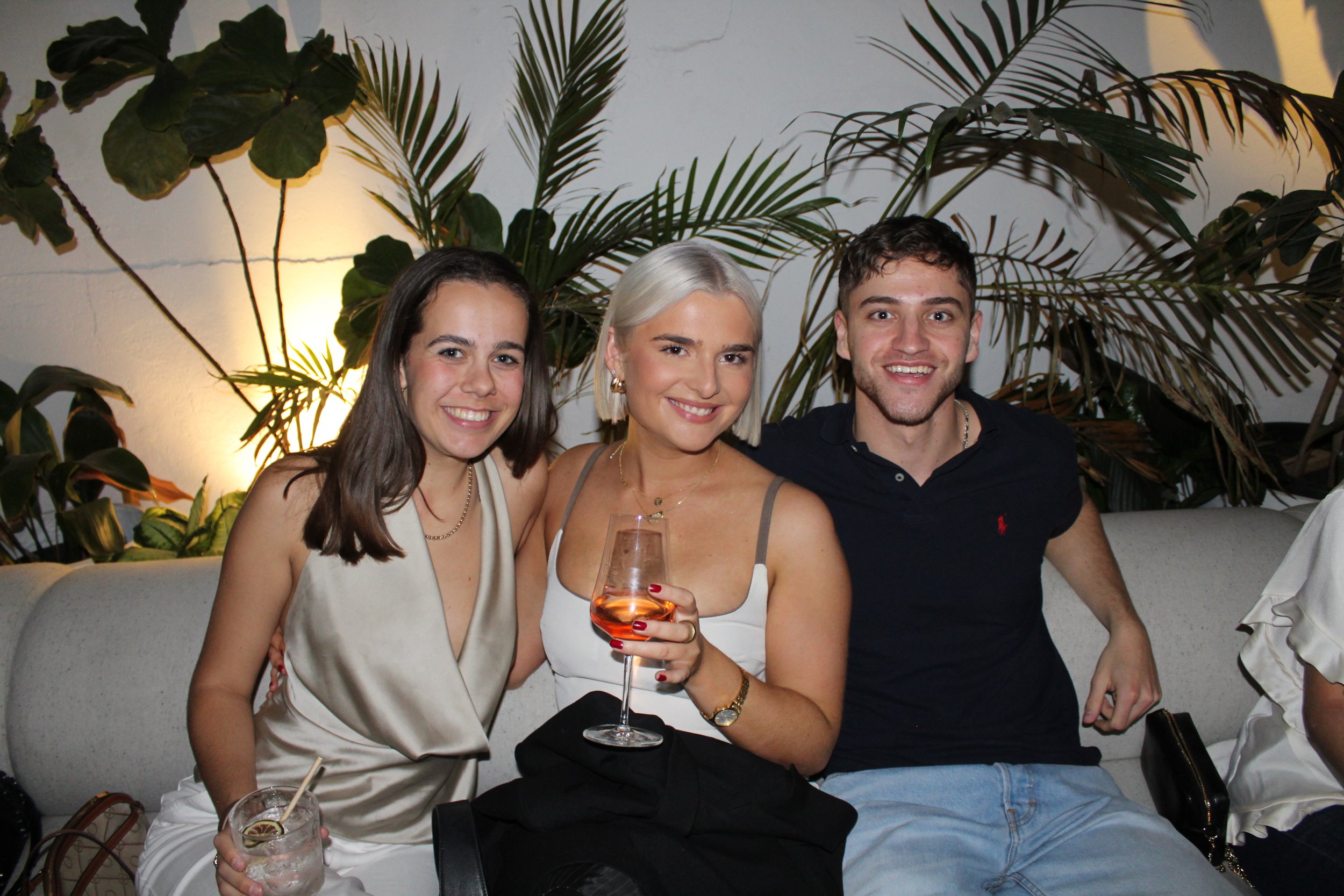 Three young adults sitting closely on a couch at a social gathering, smiling and holding drinks, with lush green plants in the background.